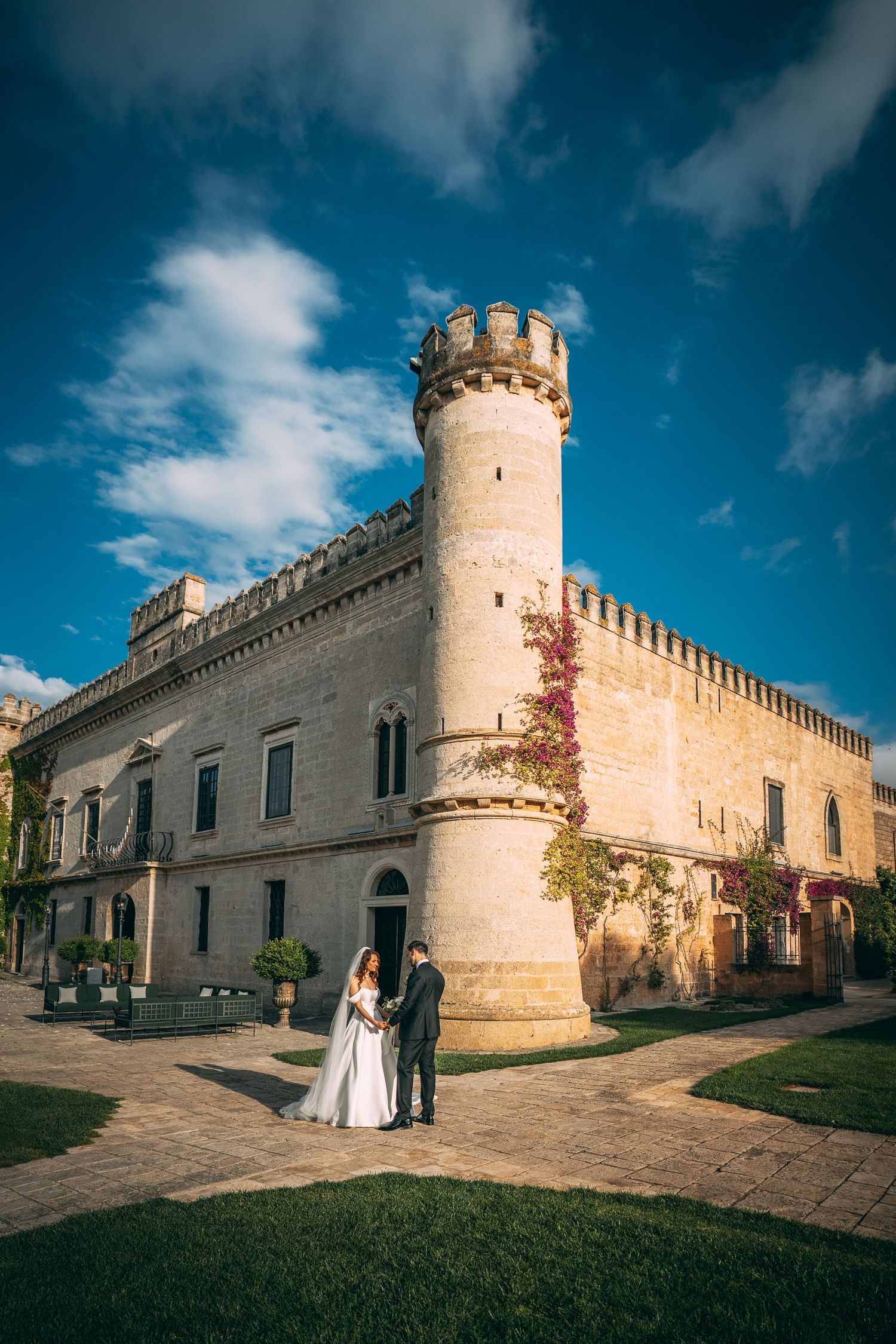Bride and groom posing in front of a historic castle with a round tower under a vibrant blue sky.