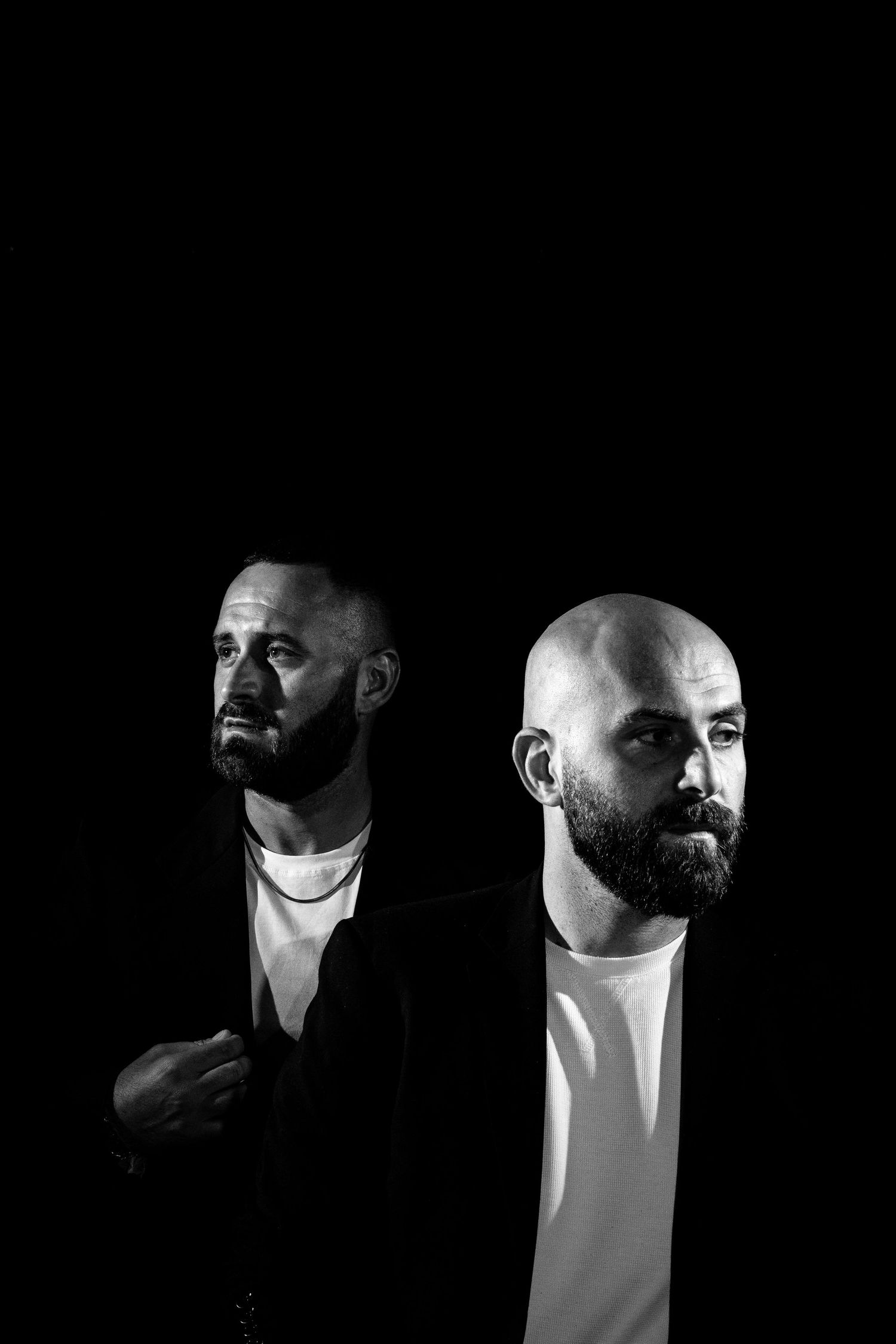 Black and white artistic portrait of two men with beards wearing white shirts against a dark background.