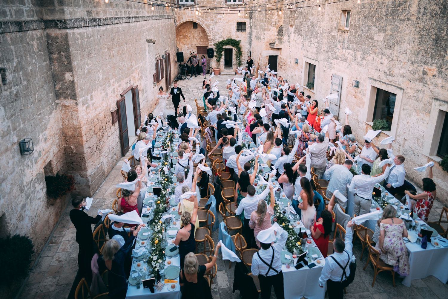 Aerial view of elegant outdoor wedding reception in historic stone courtyard with long decorated tables and guests dining.