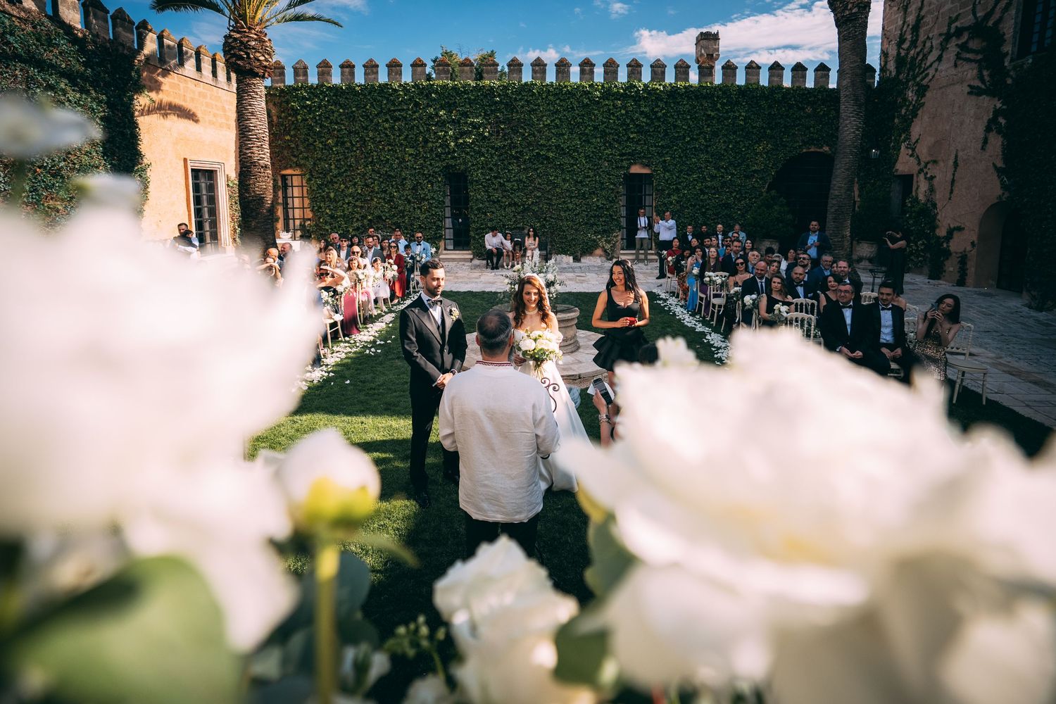 Outdoor wedding ceremony in a historic ivy-covered courtyard with white floral arrangements and seated guests.