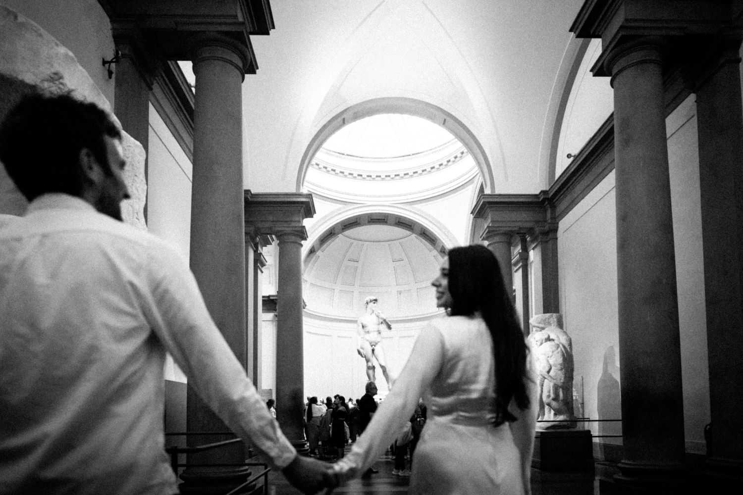 Black and white photo of a couple holding hands in the Galleria dell'Accademia, Florence, Italy.