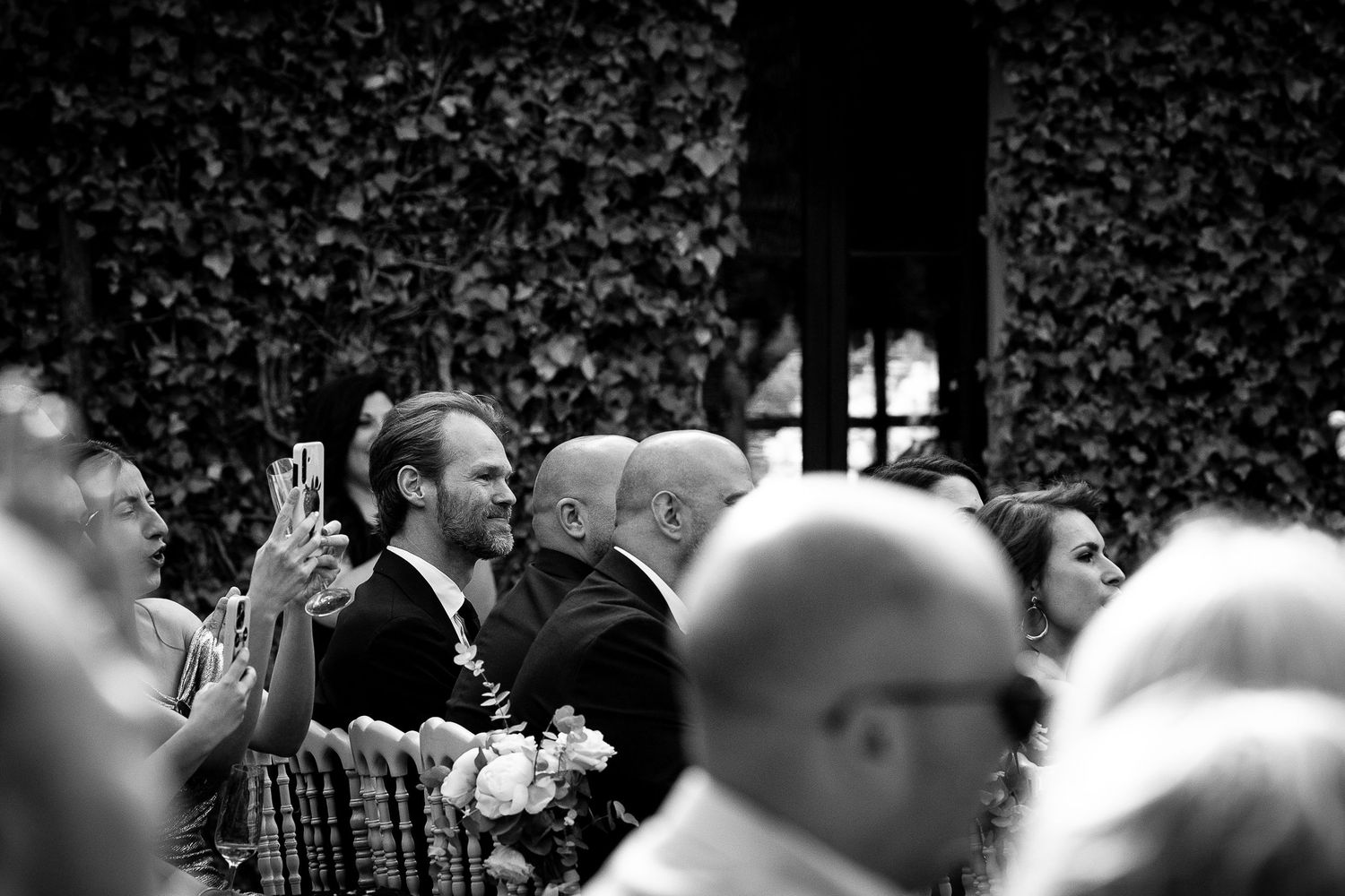 Black and white wedding guests seated outdoors, smiling and taking photos during a ceremony against an ivy-covered wall.