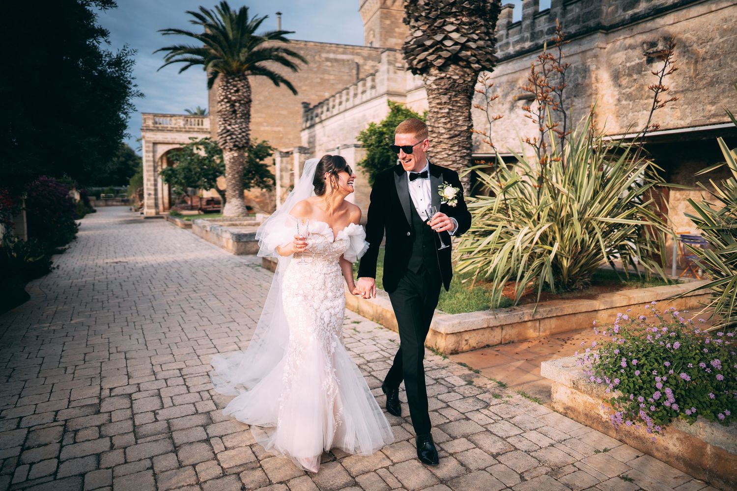 Bride and groom walking hand in hand on cobblestone path surrounded by palm trees at elegant outdoor wedding venue.