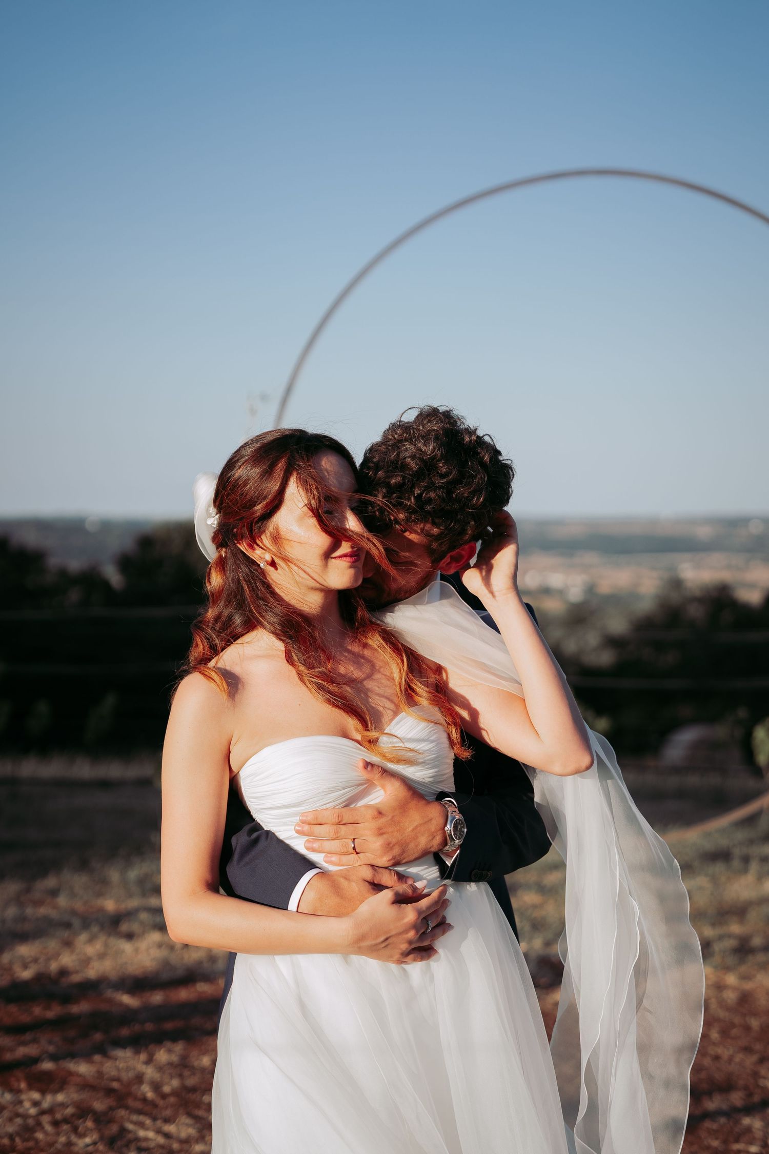 Bride and groom embracing at sunset outdoors, with a circular arch backdrop and scenic countryside views.