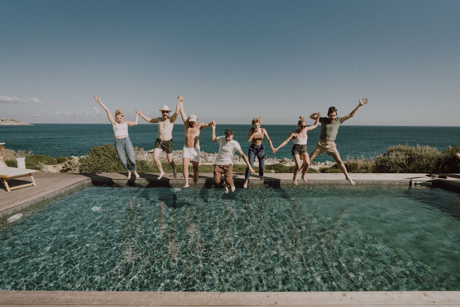 Group of friends jumping joyfully by an infinity pool overlooking the ocean on a sunny day.