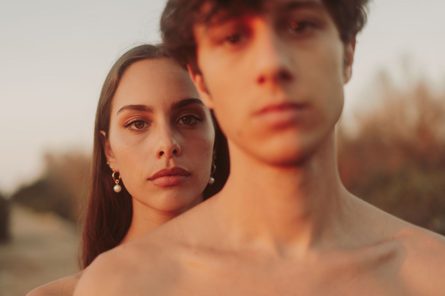 Young man and woman posing outdoors at golden hour, close-up portrait with warm sunset lighting.