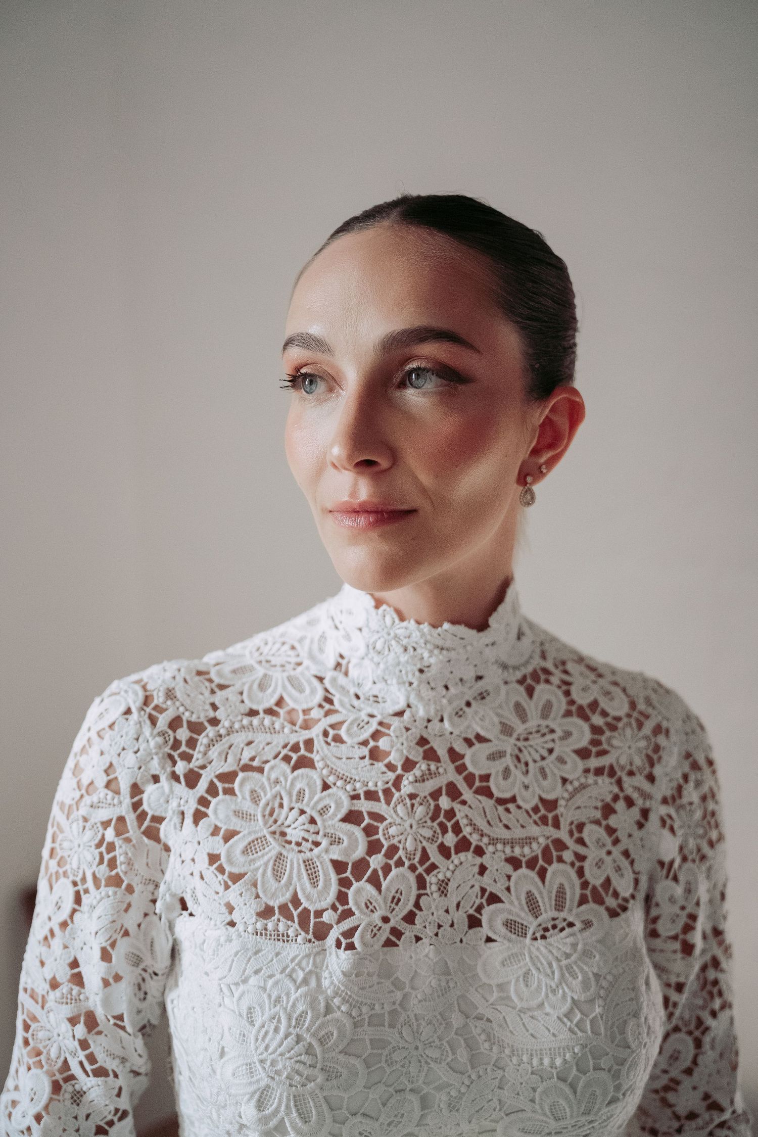 Woman in white floral lace high-neck dress with elegant updo hairstyle and pearl earring, gazing thoughtfully to the side.
