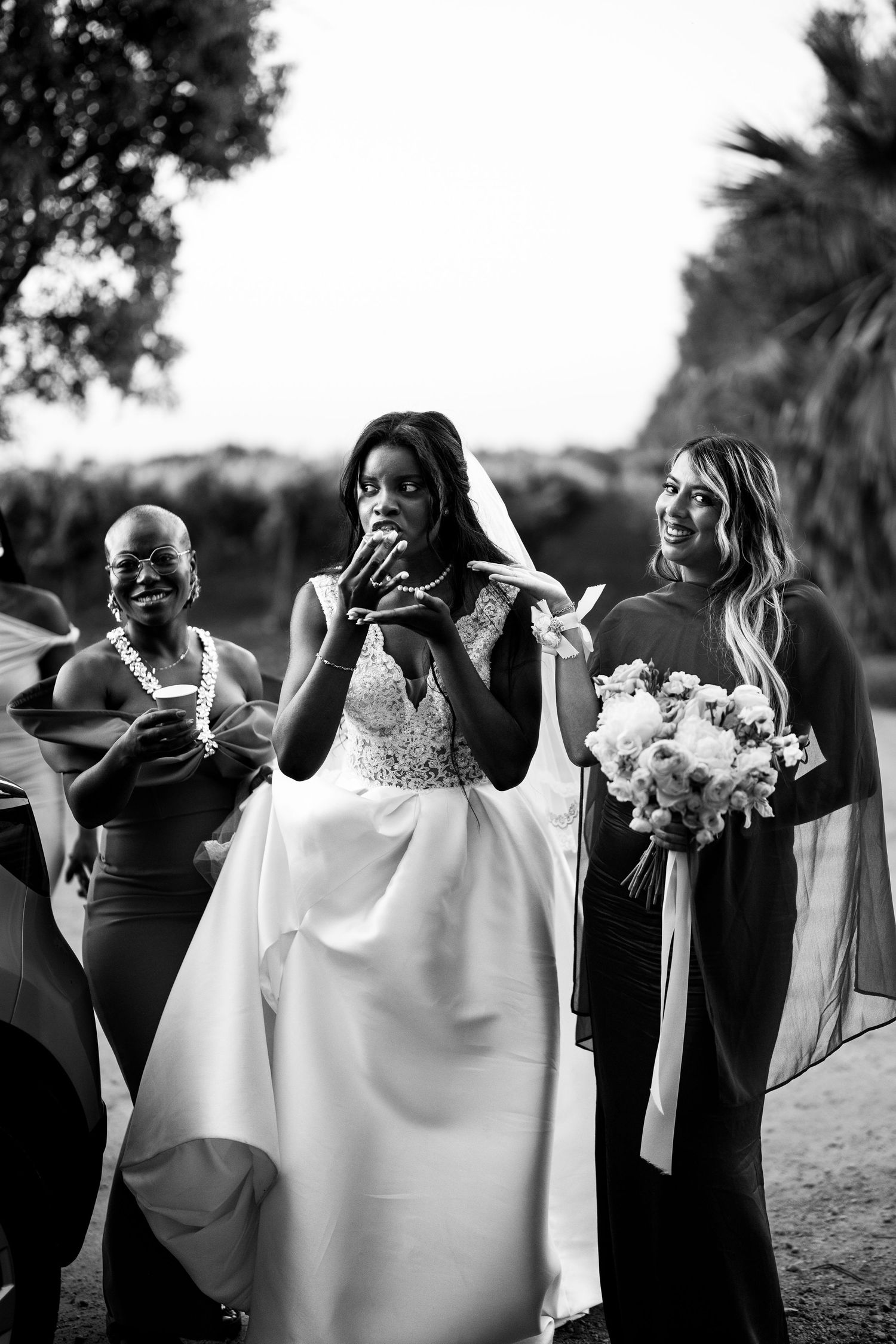 Black and white photo of a bride in lace gown with veil, flanked by bridesmaids holding bouquets at outdoor wedding.