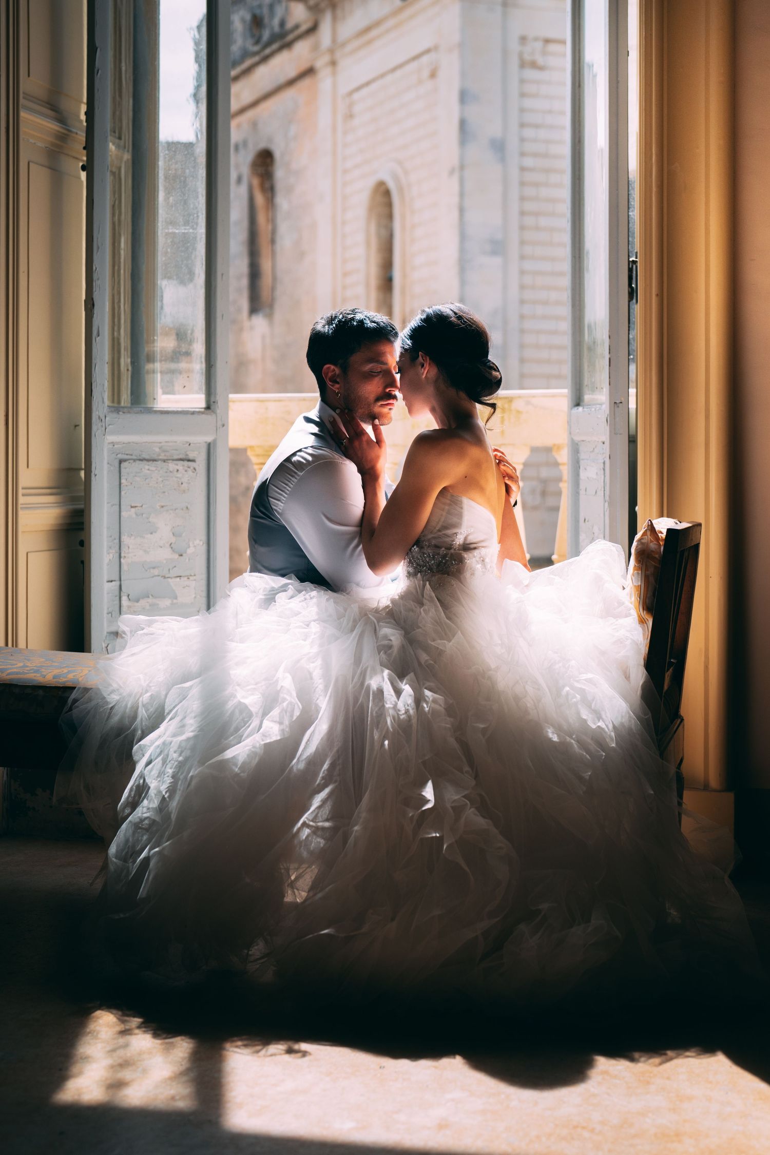 Bride in a voluminous white gown and groom share a romantic moment by sunlit vintage windows.