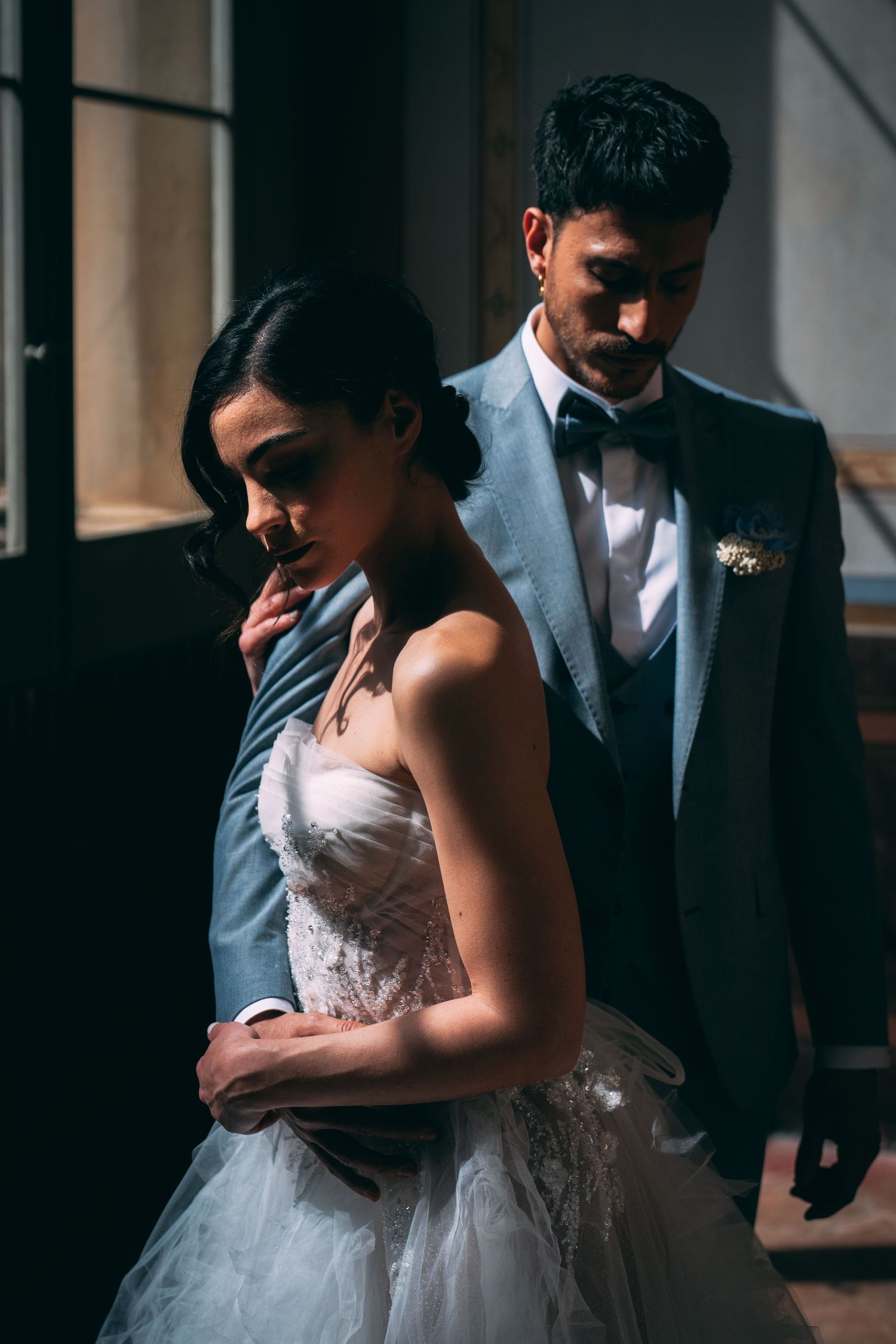 Elegant bride in white strapless gown embraced by groom in blue suit with bow tie in dramatic lighting.