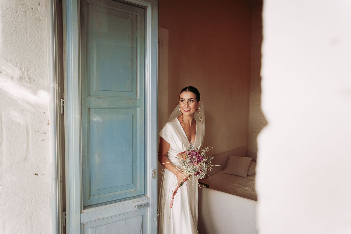 Bride in white gown holding purple bouquet stands by a blue door in a sun-drenched Mediterranean villa.