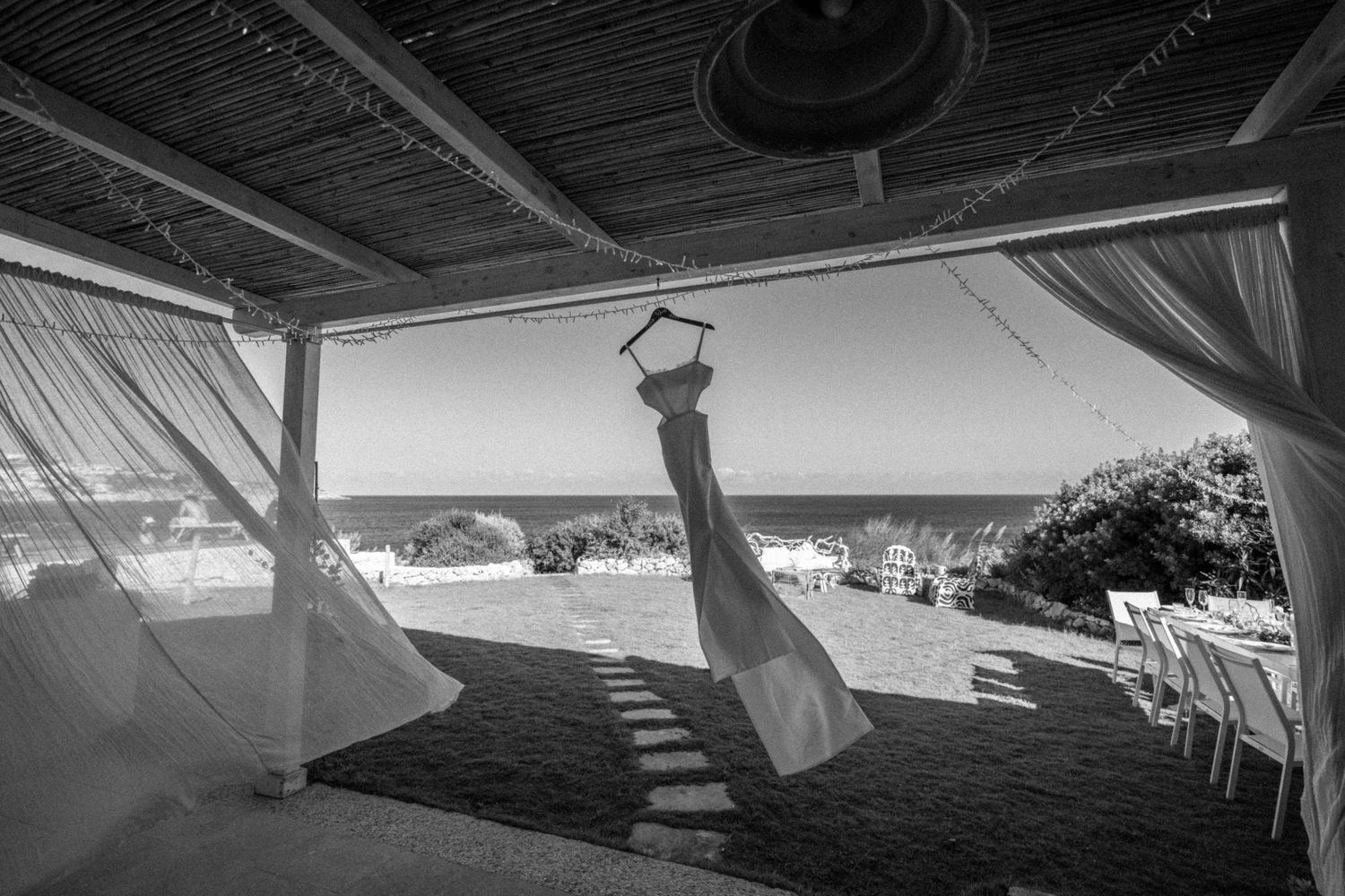 Black and white photo of a beachside wedding venue with flowing drapes, hanging decor, and ocean views.
