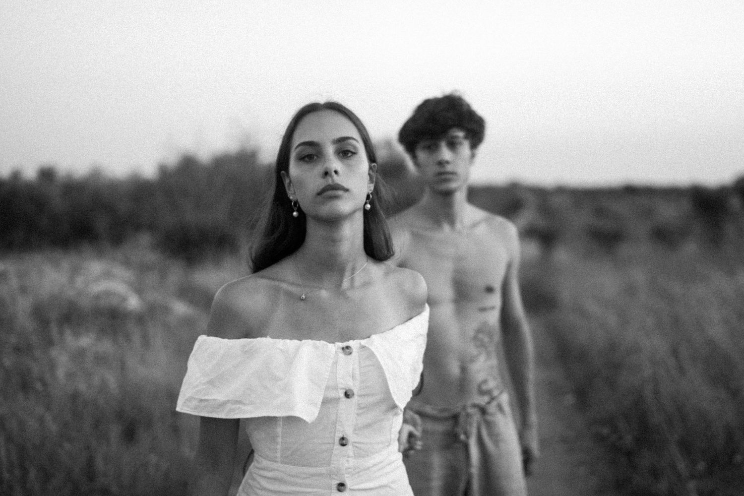 Black and white photo of a young woman in off-shoulder dress and shirtless man walking in a field.
