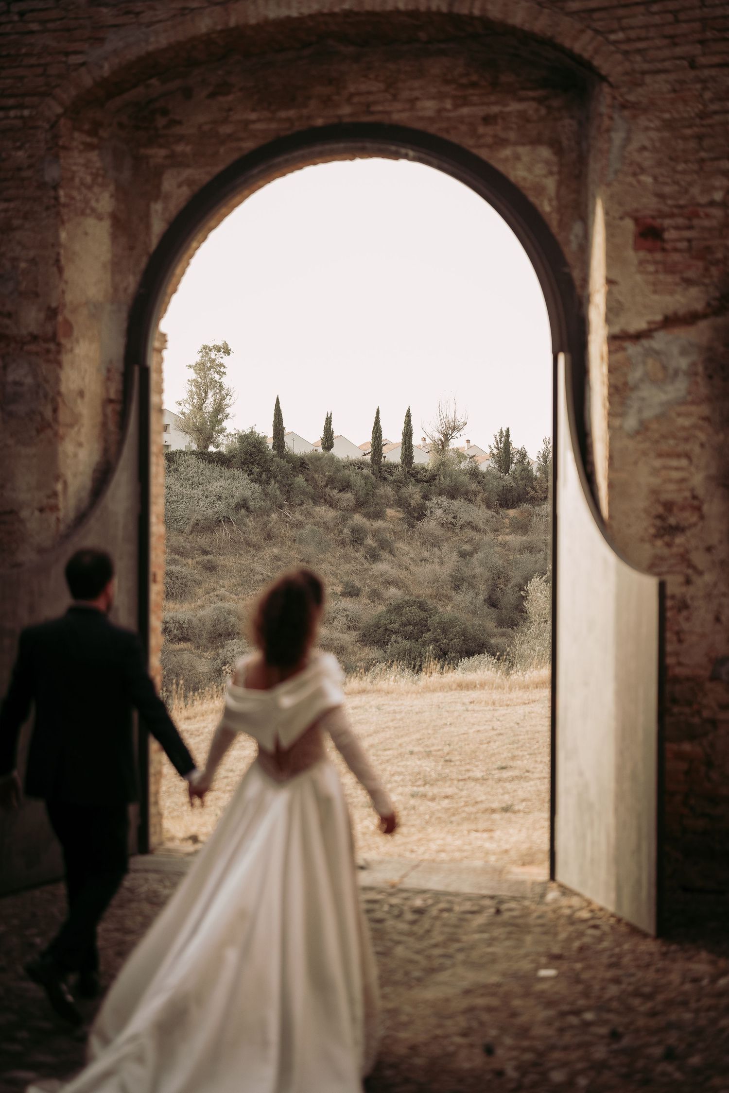 A couple in formal wedding attire walks through a rustic brick archway overlooking a scenic countryside landscape.