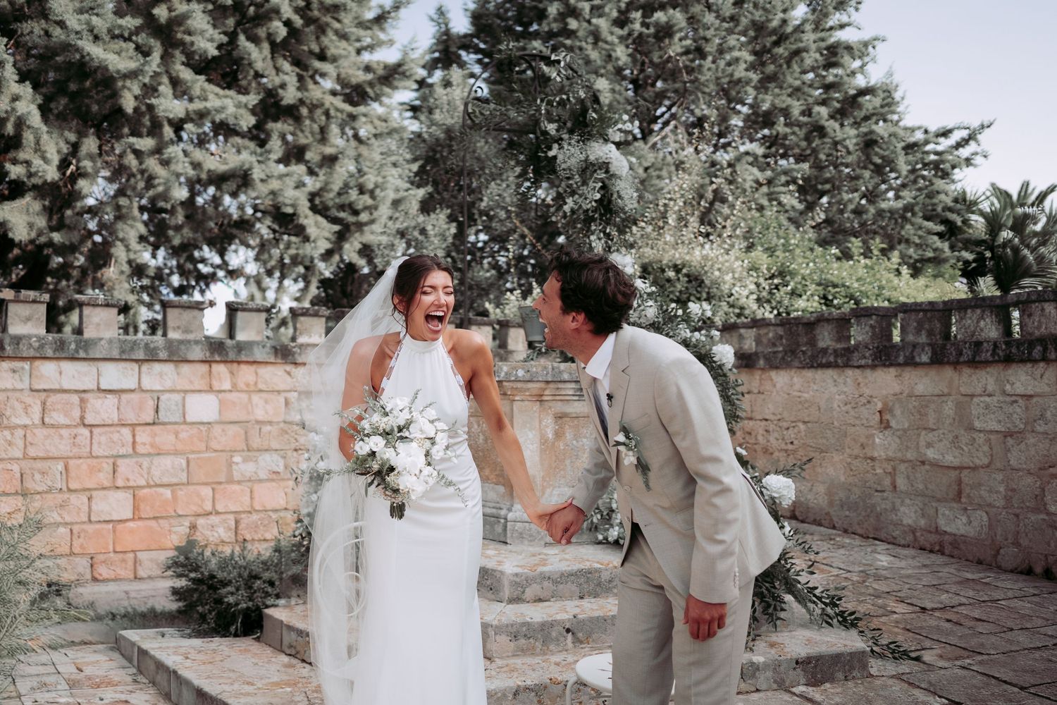 Happy bride and groom laughing together outdoors at a rustic stone venue, bride holding white bouquet in wedding dress.