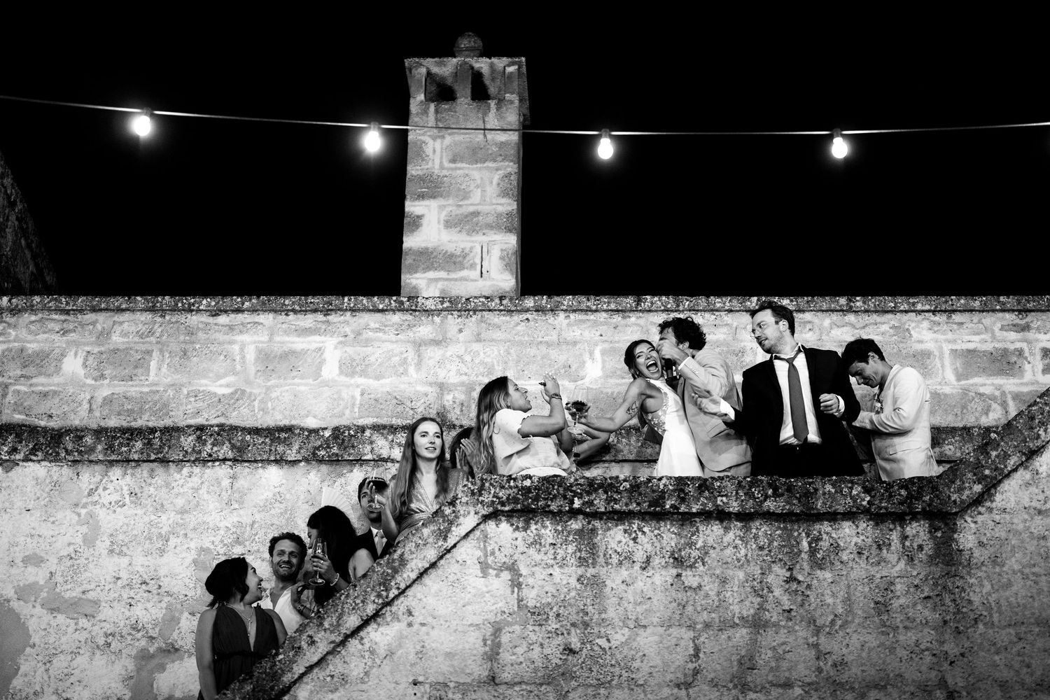 A black and white photo montage shows people enjoying a nighttime outdoor gathering near stone walls with string lights.