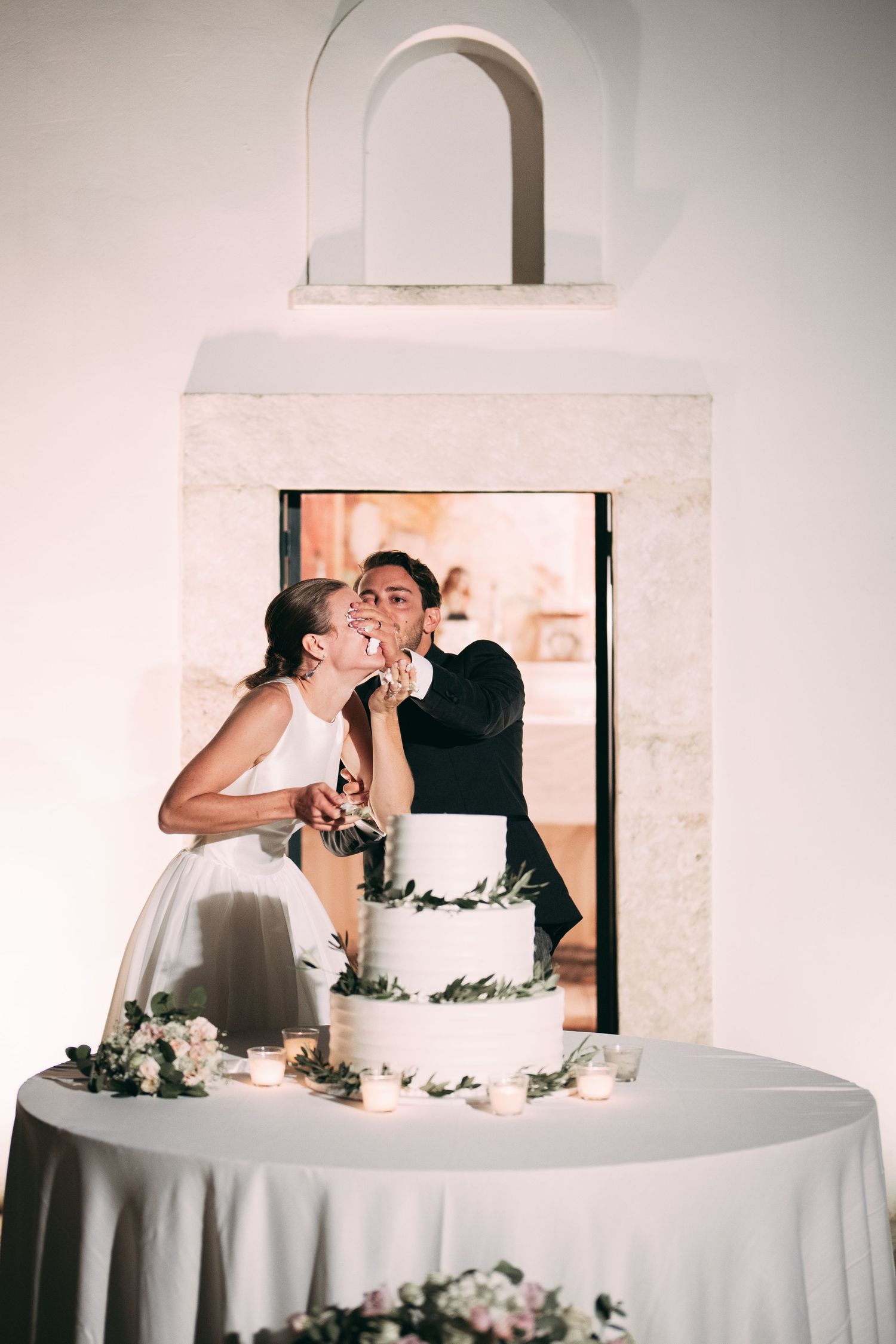 Bride and groom kissing while cutting their white tiered wedding cake decorated with greenery at their reception.