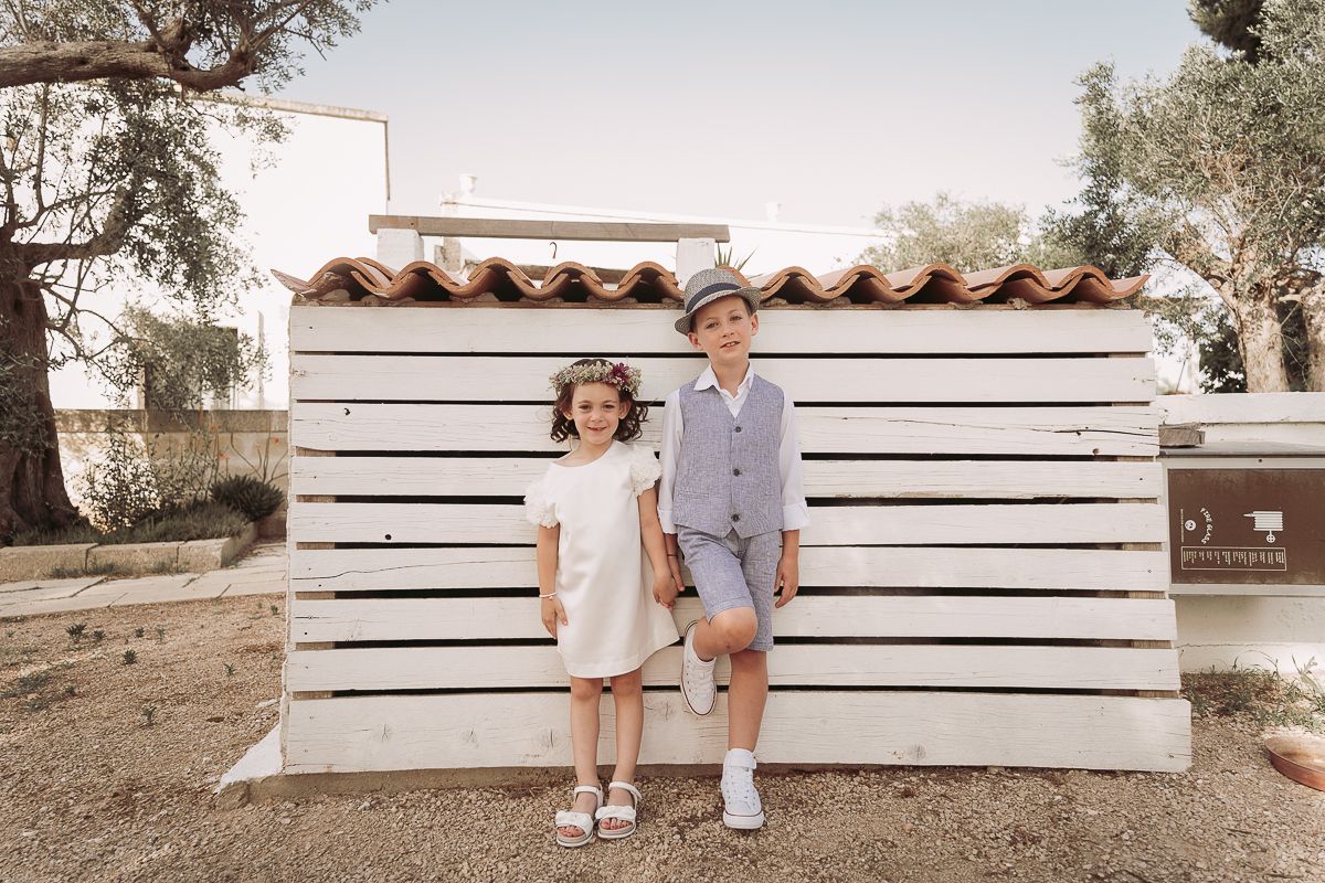 Two children in elegant outfits pose in front of a white wooden structure outdoors.
