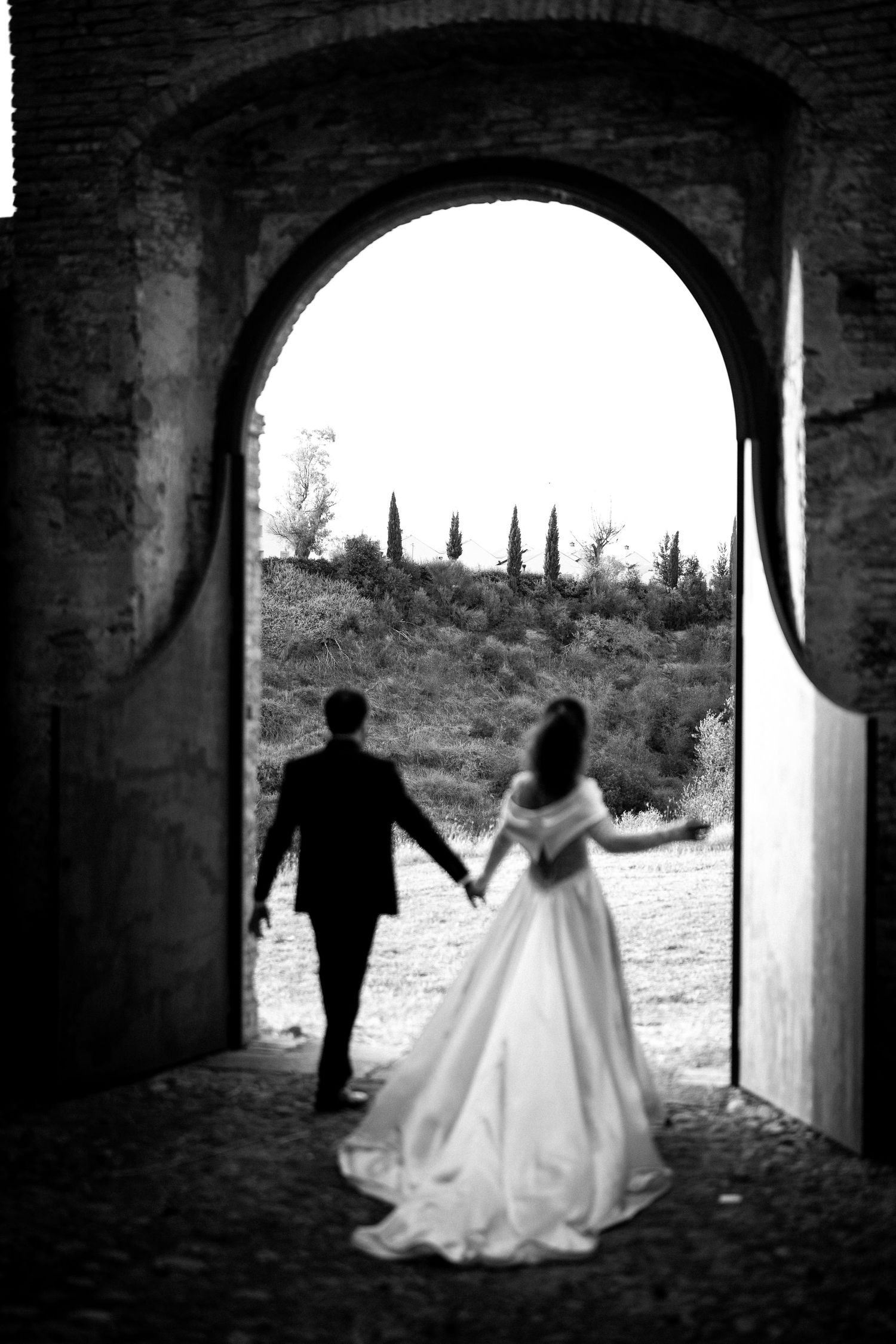 Black and white photo of bride and groom holding hands walking through stone archway toward scenic countryside.