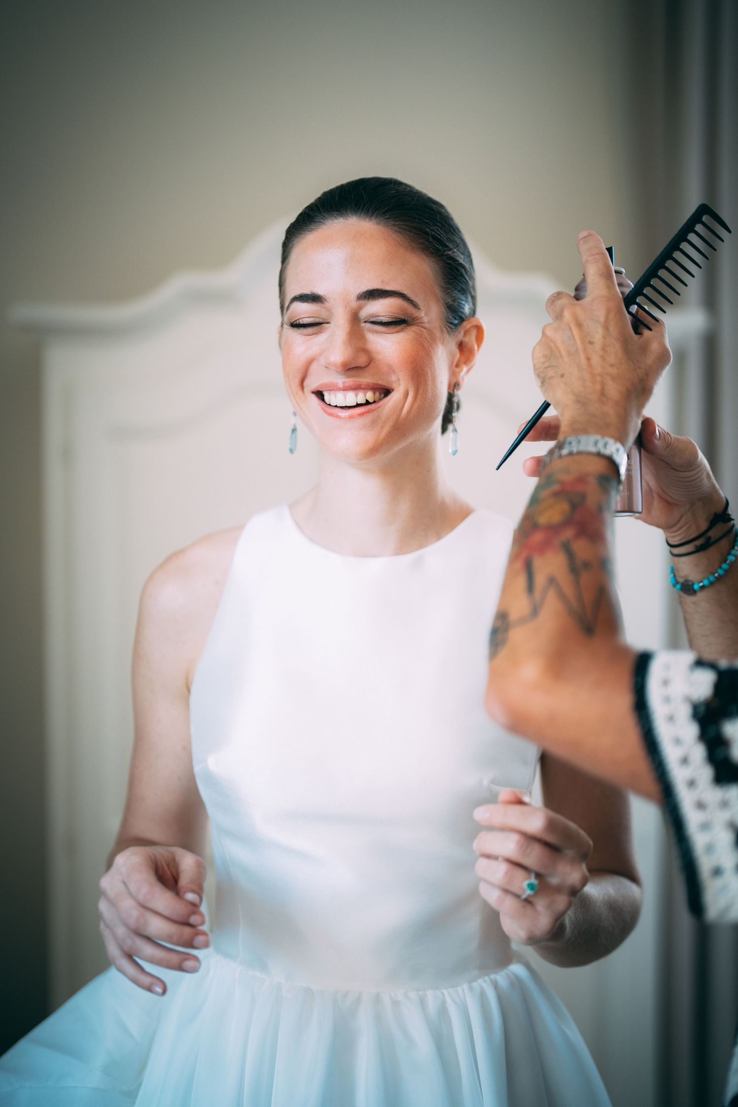 Smiling bride in white dress getting hair styled by tattooed hairstylist holding comb and scissors.