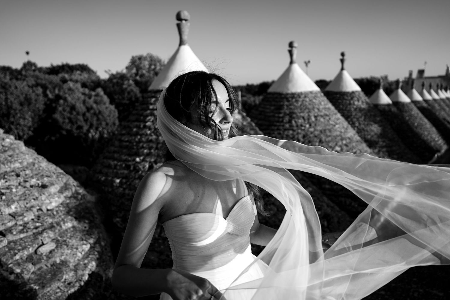 Black and white photo of a bride with flowing veil blowing in wind, standing among traditional trulli stone huts.