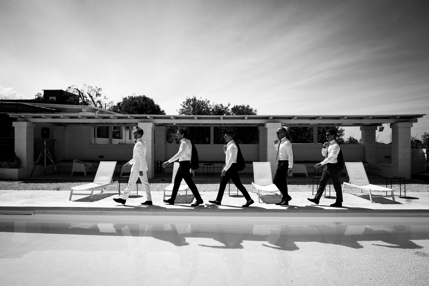 Five men in formal attire walk in a line beside a pool at a mid-century modern home in black and white.