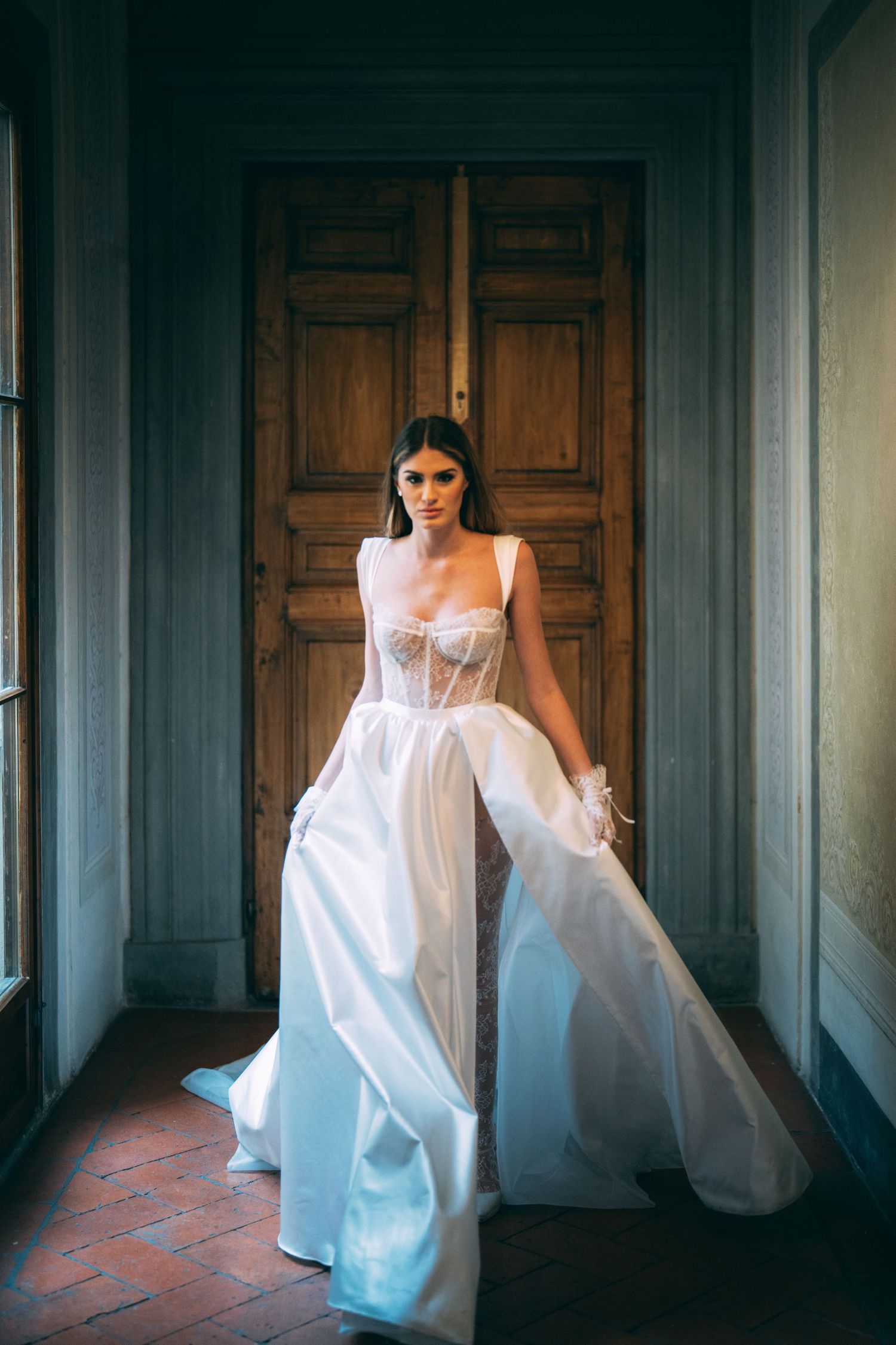 Bride in a white corset wedding gown with flowing skirt stands before wooden doors in an elegant venue.