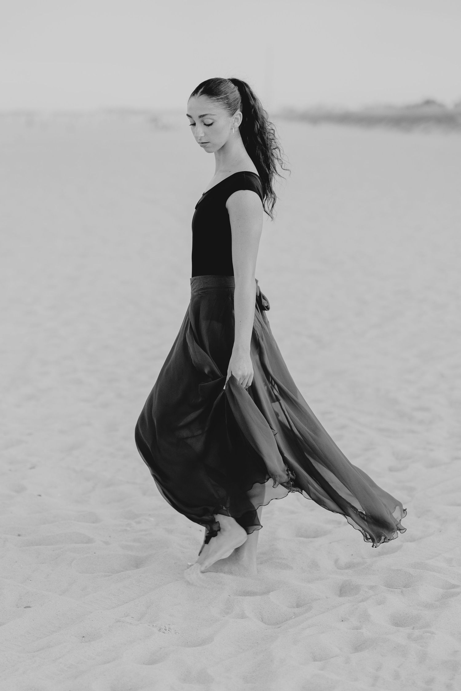 Graceful dancer in flowing dark skirt and black top walks barefoot on sandy beach in elegant black and white photo.