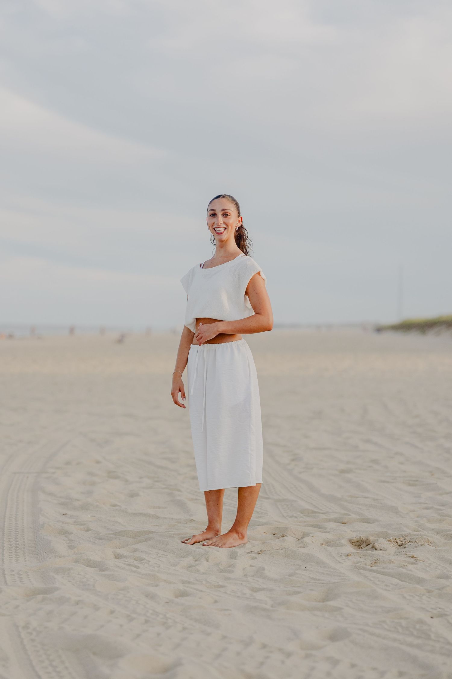 Woman in white crop top and midi skirt standing barefoot on sandy beach smiling.