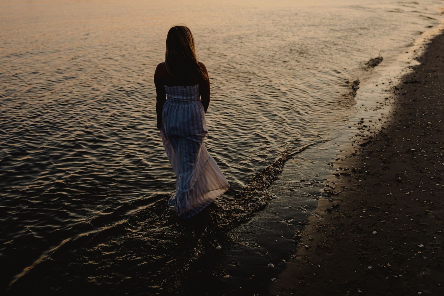 A woman in a flowing white dress wades through shallow ocean water at sunset on a serene beach.