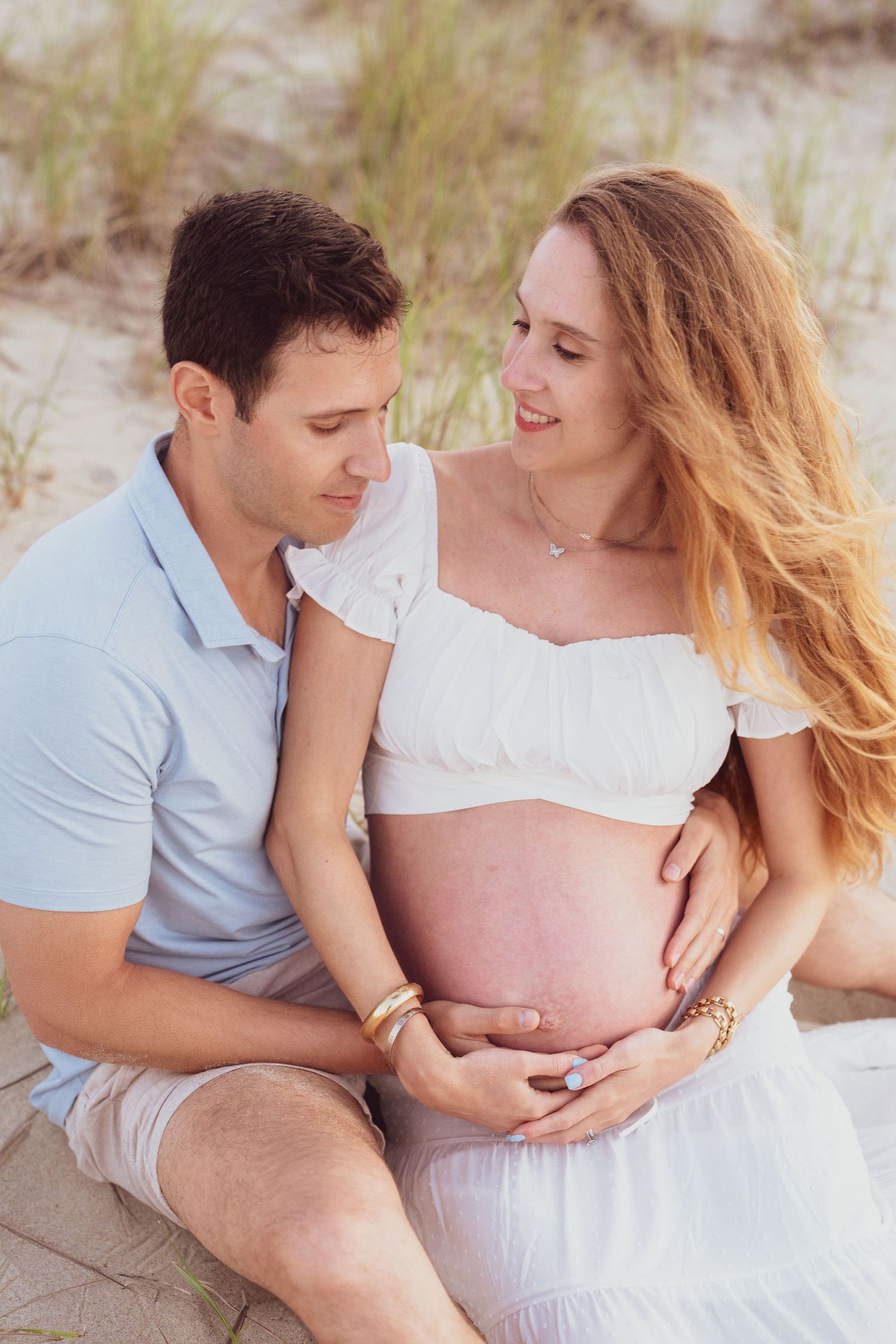 A couple in coordinating outfits shares intimate moments on a sandy beach with sea grass.