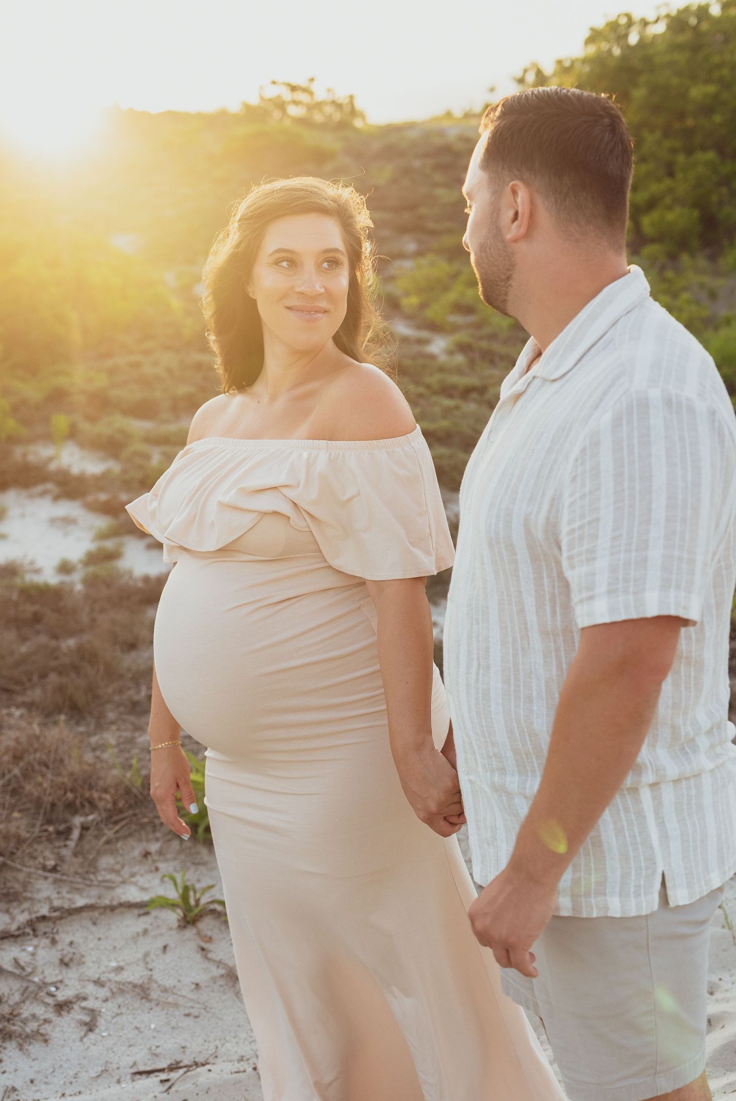 An expectant couple shares a tender moment at sunset on a sandy beach.