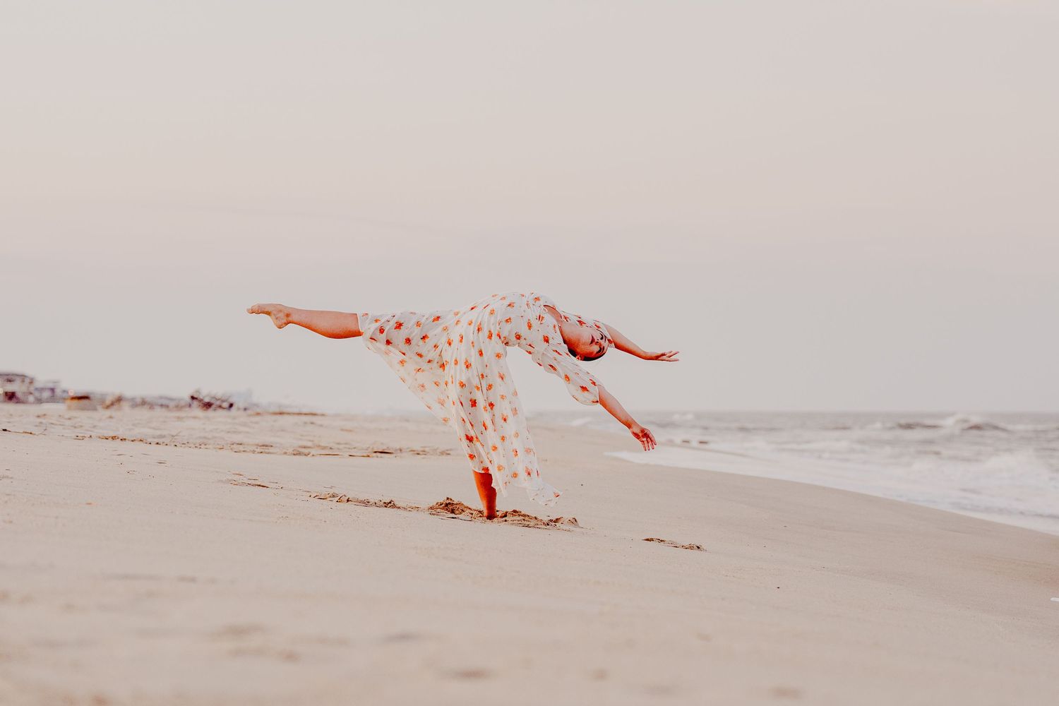 Woman in white floral dress doing a cartwheel on a sandy beach at sunset near the ocean.