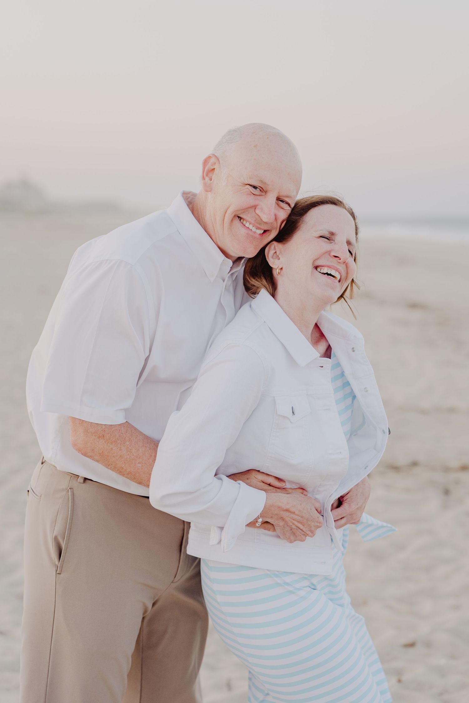 Happy couple embracing and laughing on a sandy beach at sunset, wearing white and khaki casual outfits.