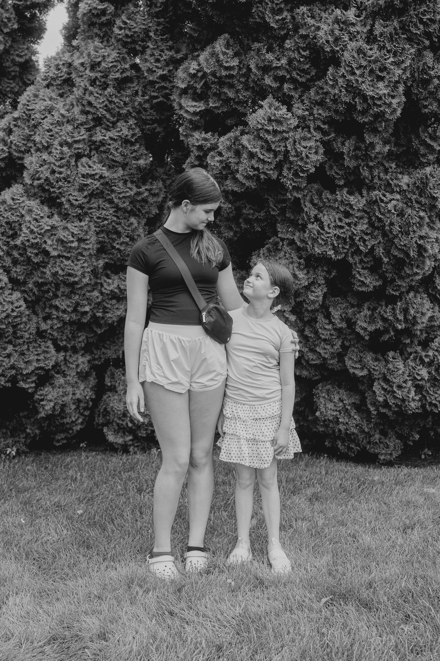 Black and white photo of older girl and young child standing together on grass in front of evergreen trees.