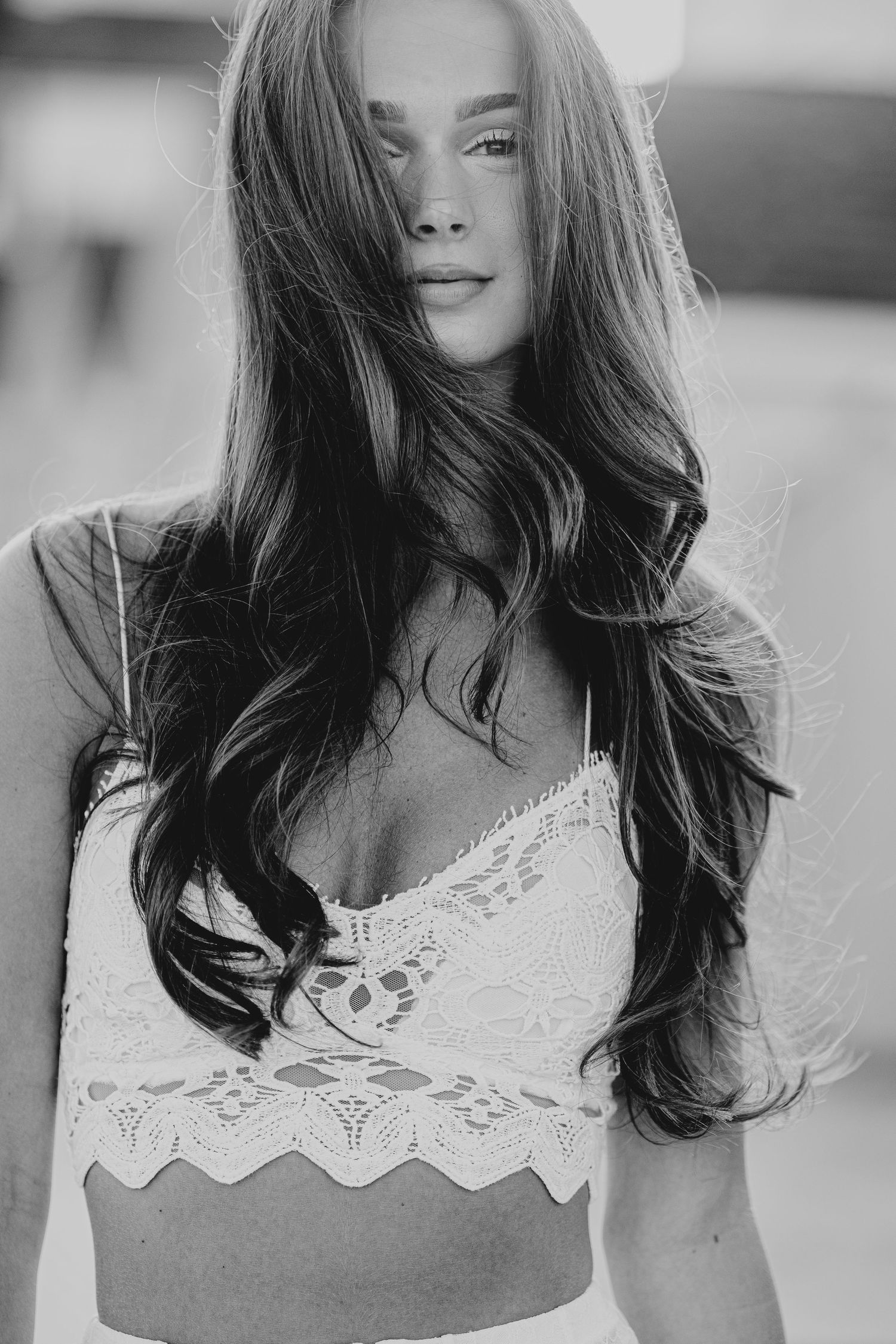 Black and white portrait of a young woman with long wavy hair wearing a white lace bralette top.
