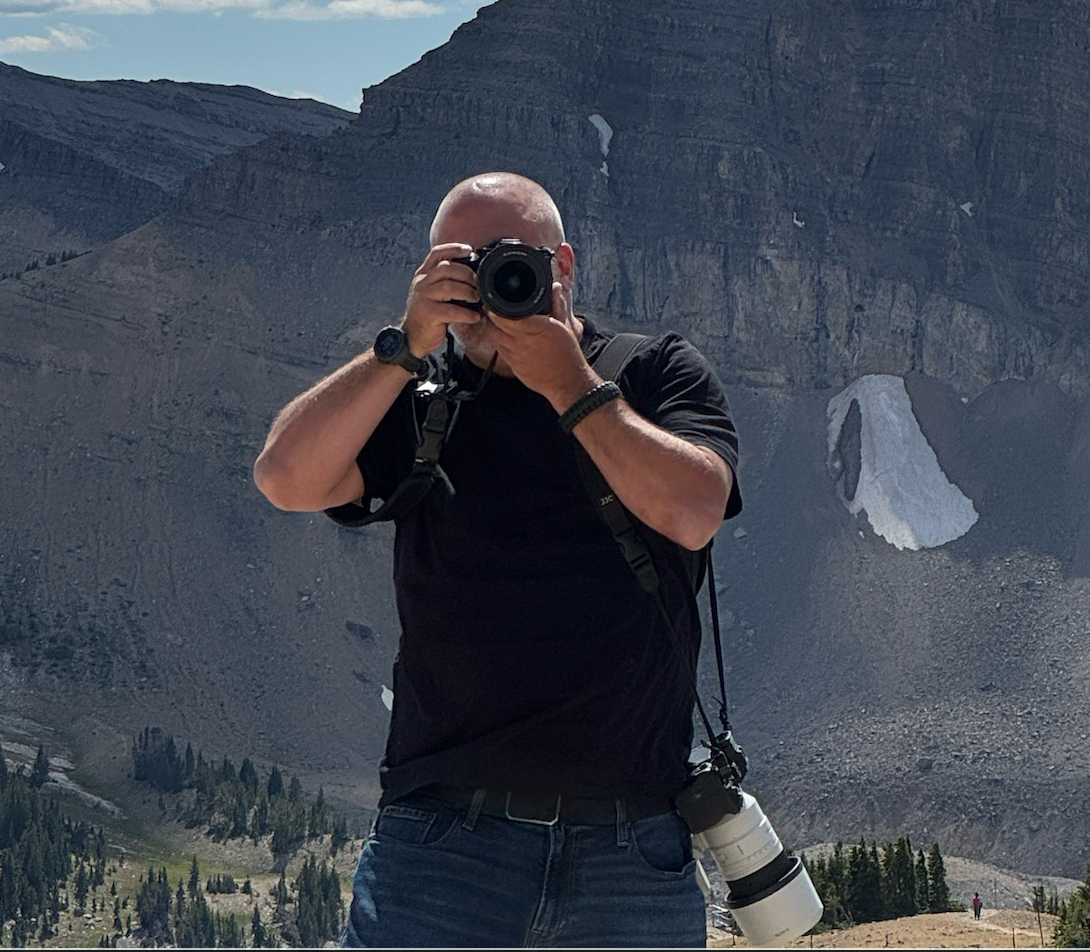 Photographer capturing mountain landscape with DSLR camera, wearing black shirt, with scenic rocky peaks and forest in background.