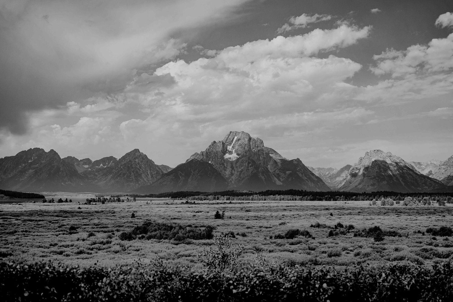 Panoramic black and white landscape of Grand Teton mountain range with dramatic cloudy sky and sweeping valley below.