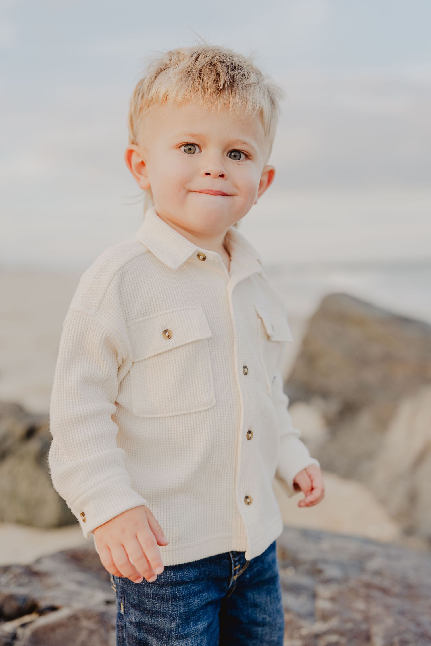 A cute blond toddler boy in a cream button-up shirt smiles near rocky shore.