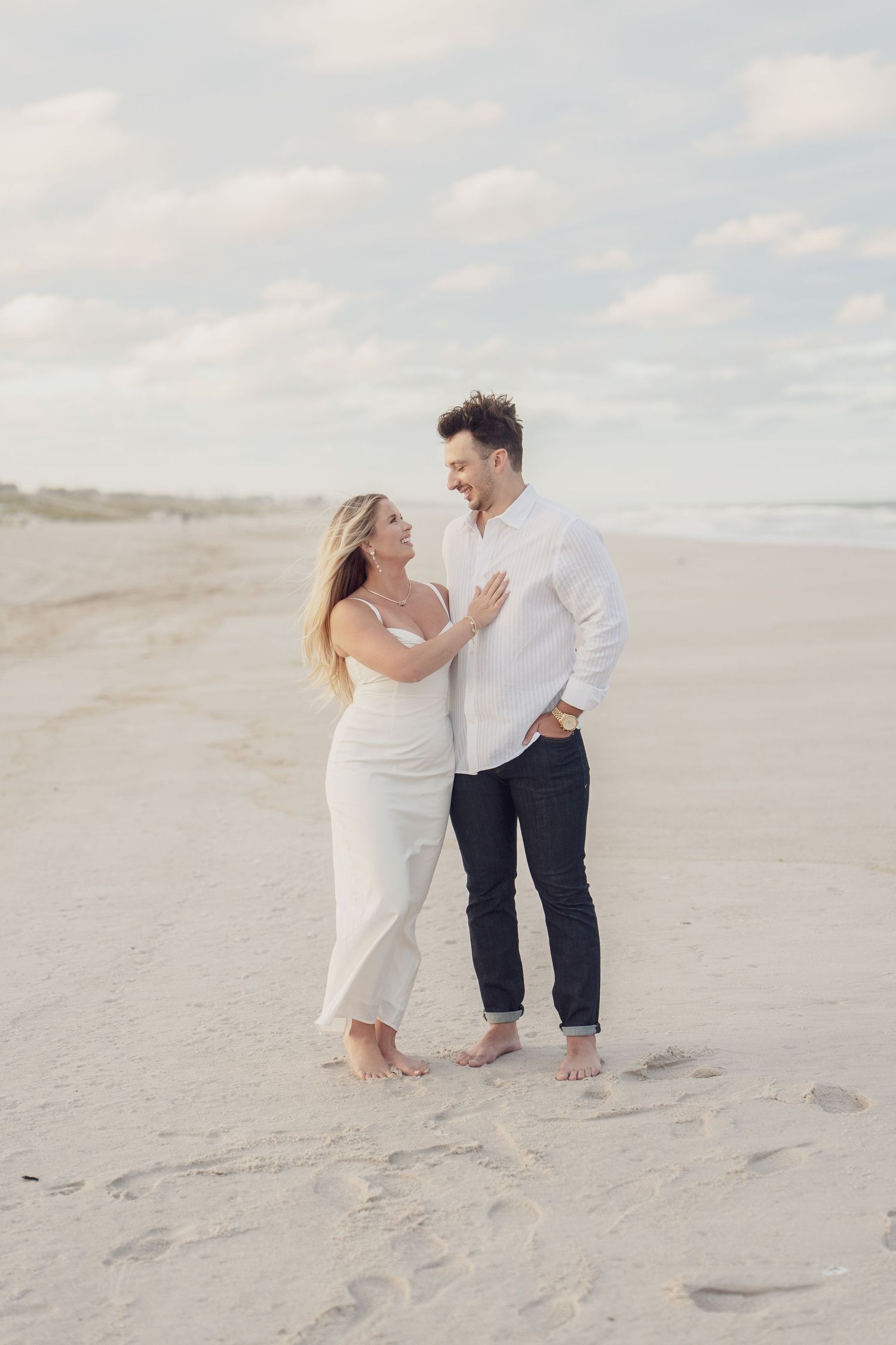 Playful couple in white clothing dancing on sandy beach with cloudy sky backdrop.
