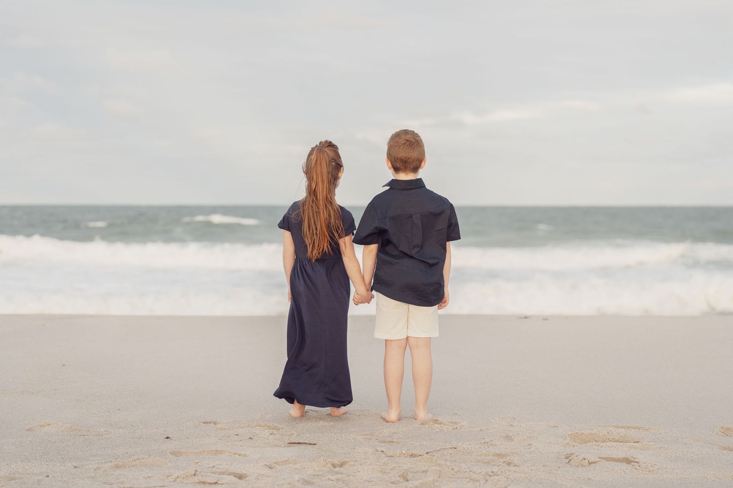 Two people stand together on a sandy beach looking out at the ocean waves under an overcast sky.