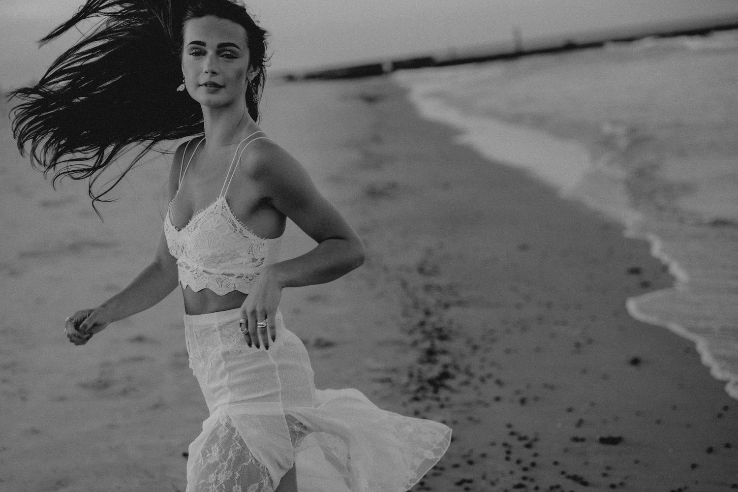 Black and white photo of a woman in a white dress running along a beach with wind blowing her hair.