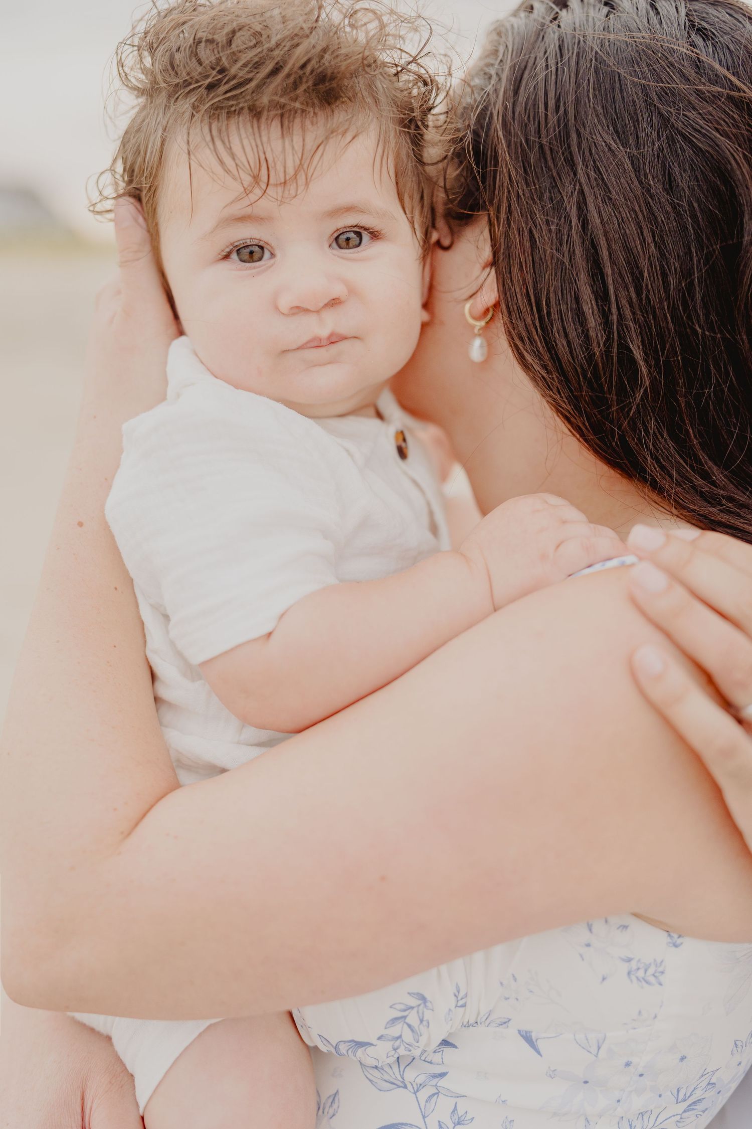 A mother tenderly kisses her curly-haired baby boy dressed in white, captured in a soft, dreamy outdoor portrait.