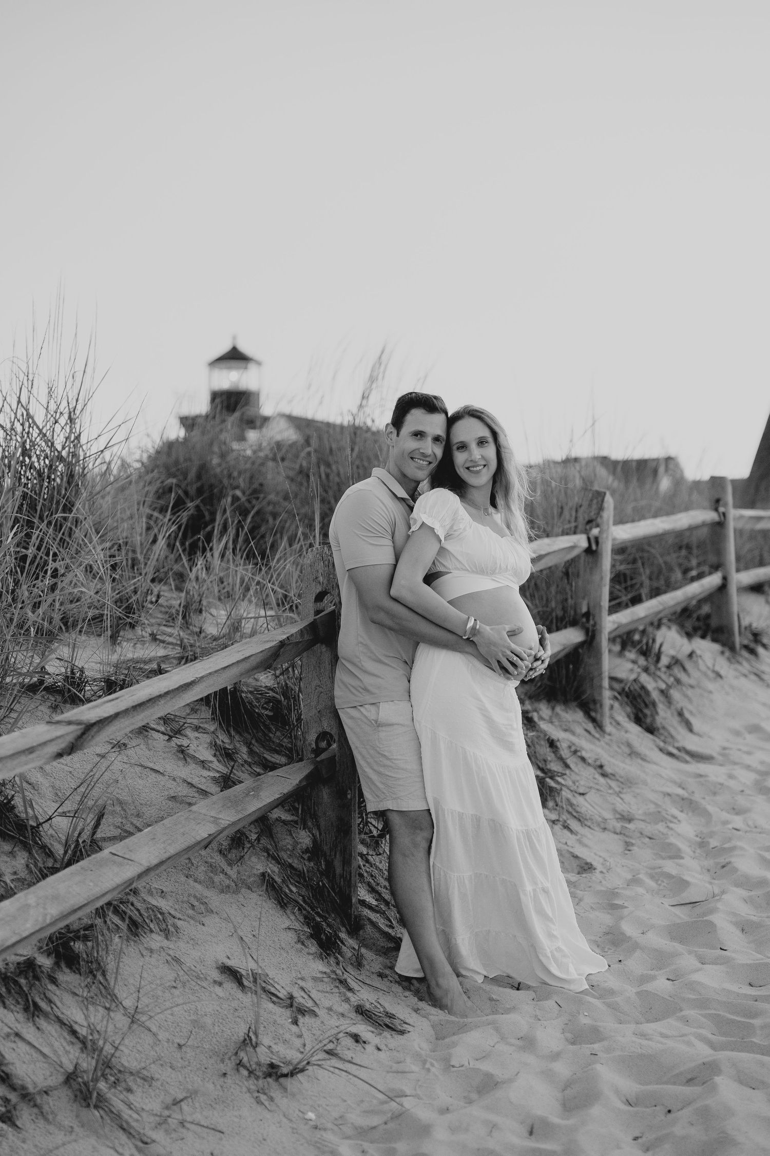 A couple poses together on sandy dunes with a wooden fence in black and white.