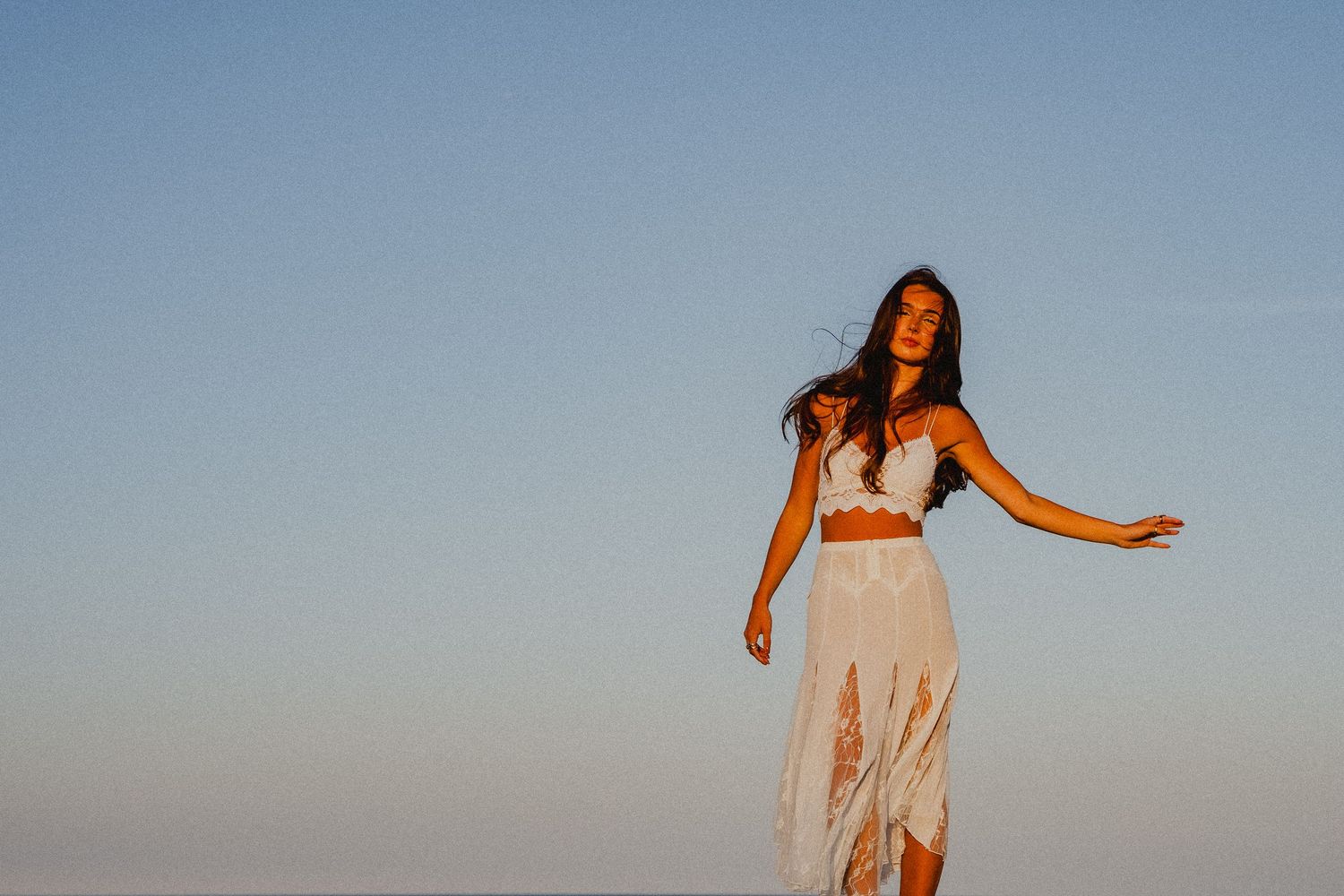 Woman in white bohemian crop top and maxi skirt posing outdoors against a clear blue sky at sunset.