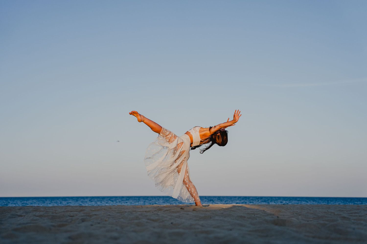 Woman in flowy white dress dancing gracefully on a sandy beach at sunset with blue sky and ocean backdrop.