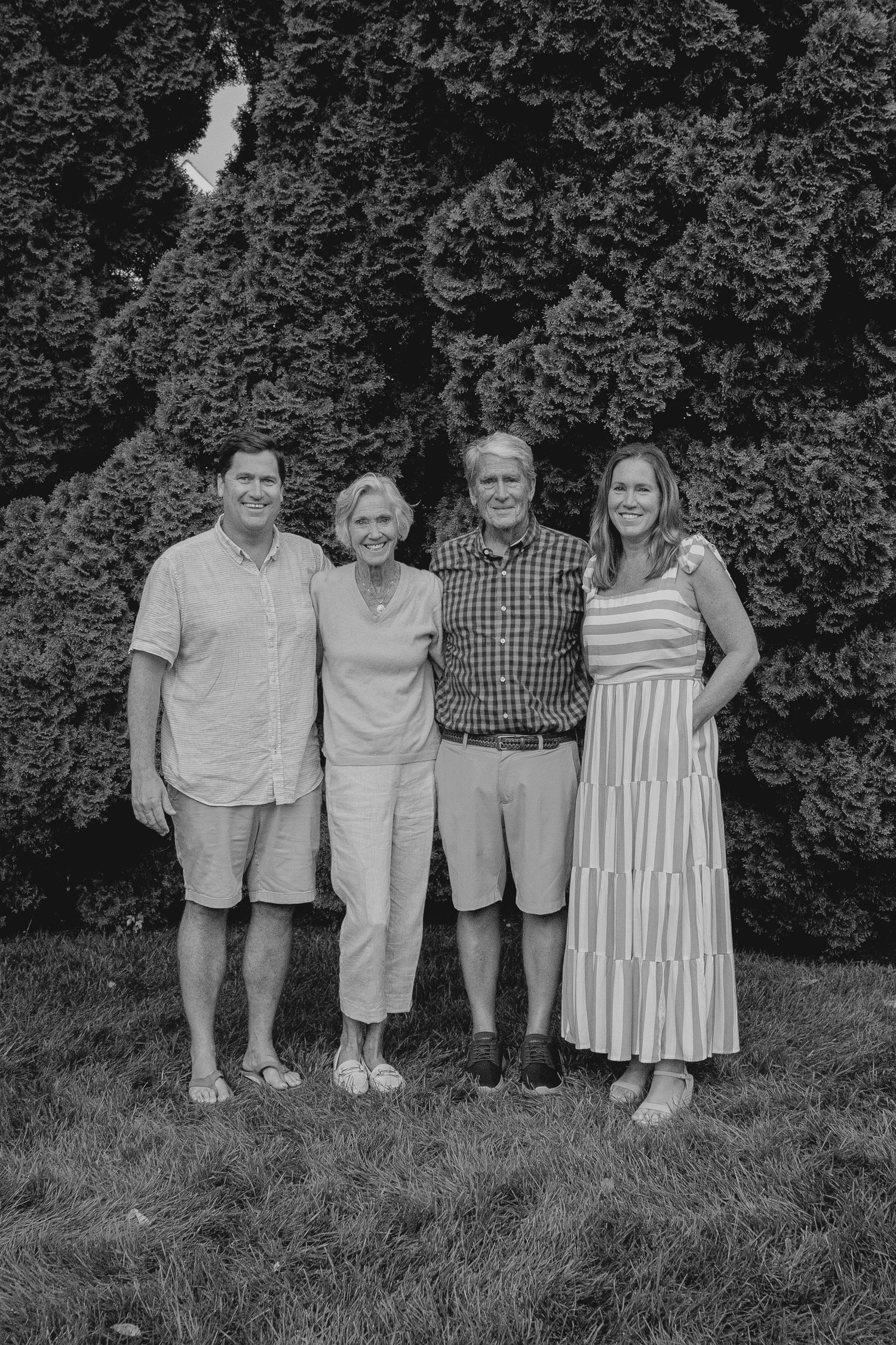 Black and white family portrait of four adults smiling outdoors in front of lush evergreen trees.