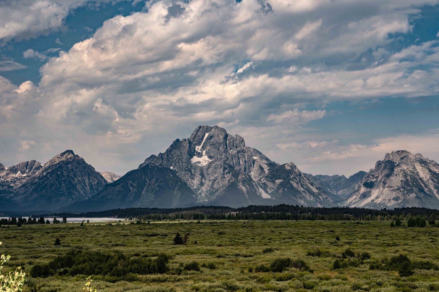 Majestic Grand Teton peaks rise above green meadows under dramatic cloudy sky.