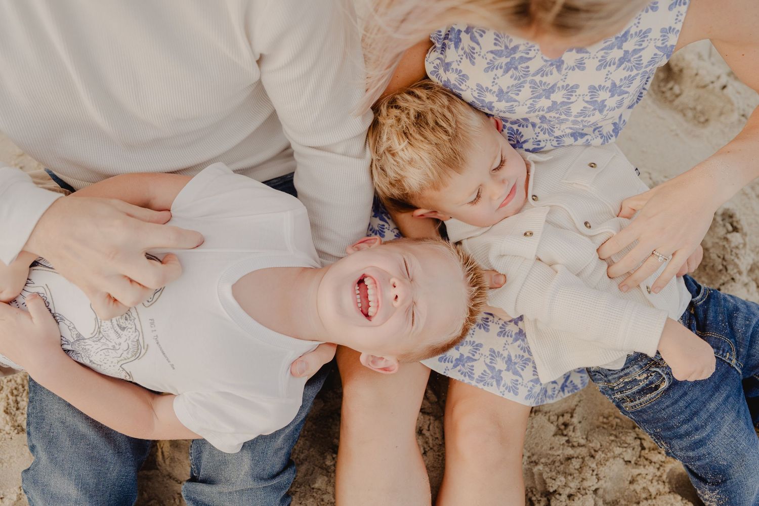 Two young children laughing and playing on sandy beach with parents during family photo session.