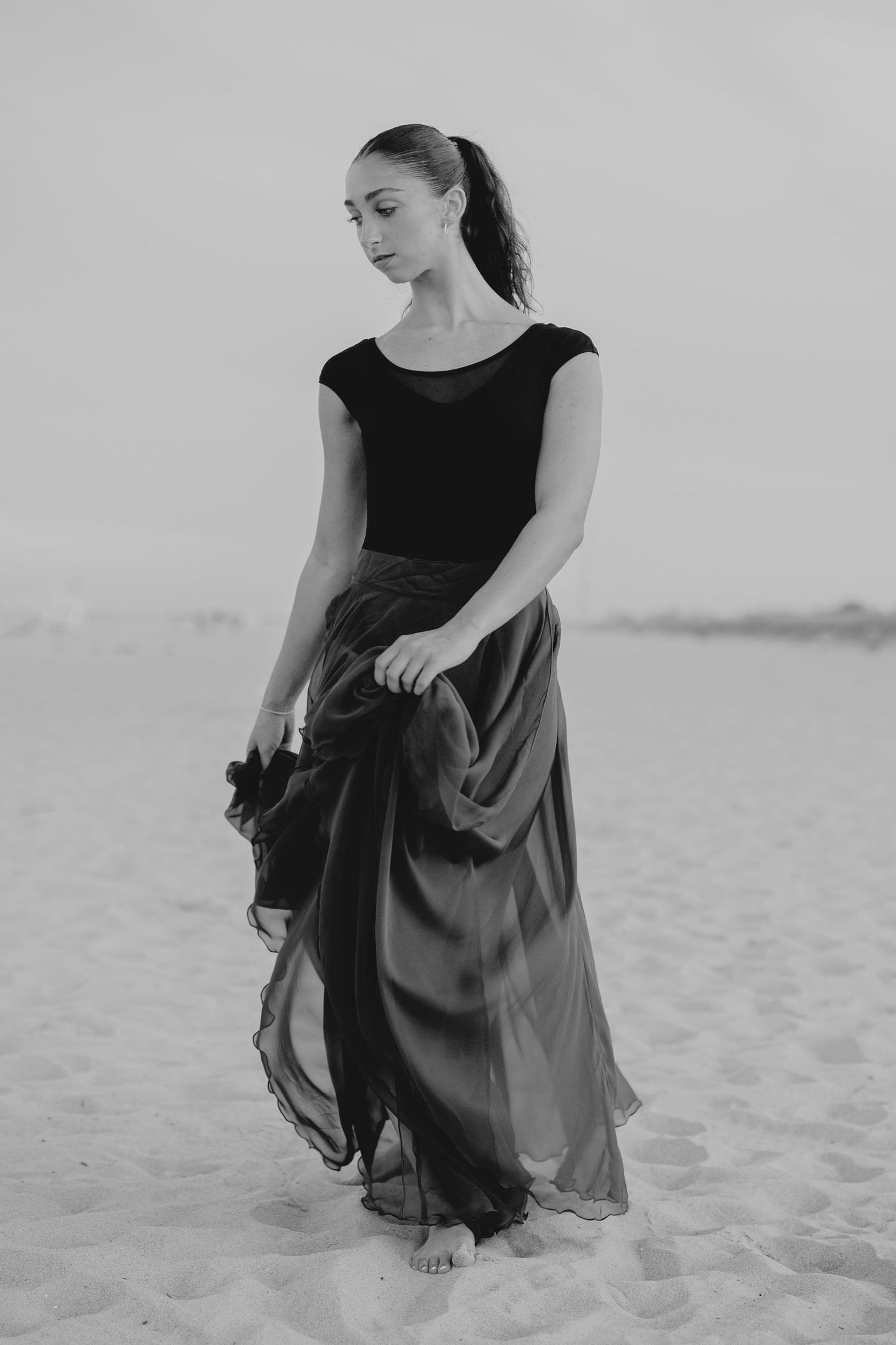 Black and white photo of a woman in a flowing dark skirt and black top standing barefoot on sandy beach.